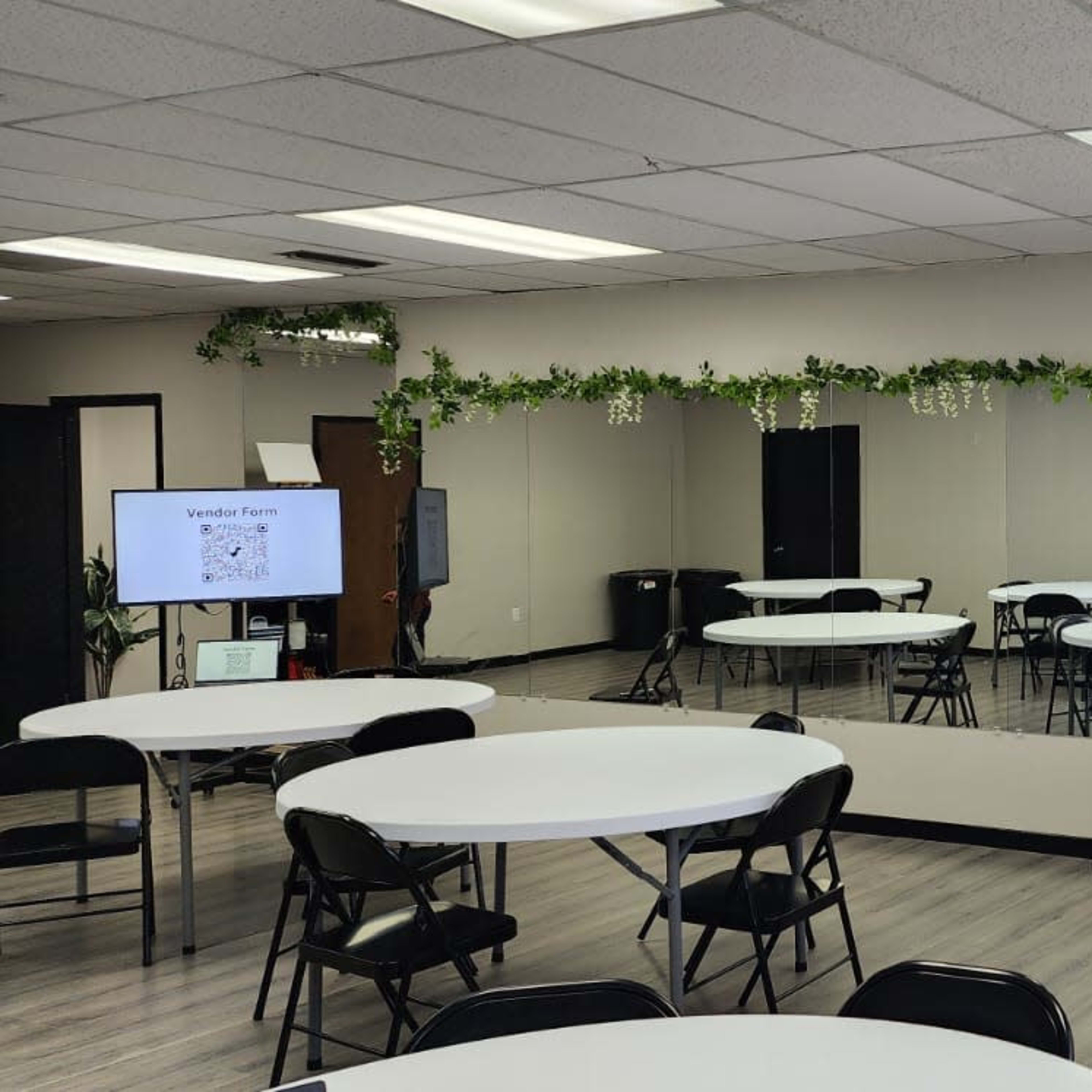 A spacious room with several round tables and black chairs arranged around them, and a long mirror on one wall with decorative greenery at the top.