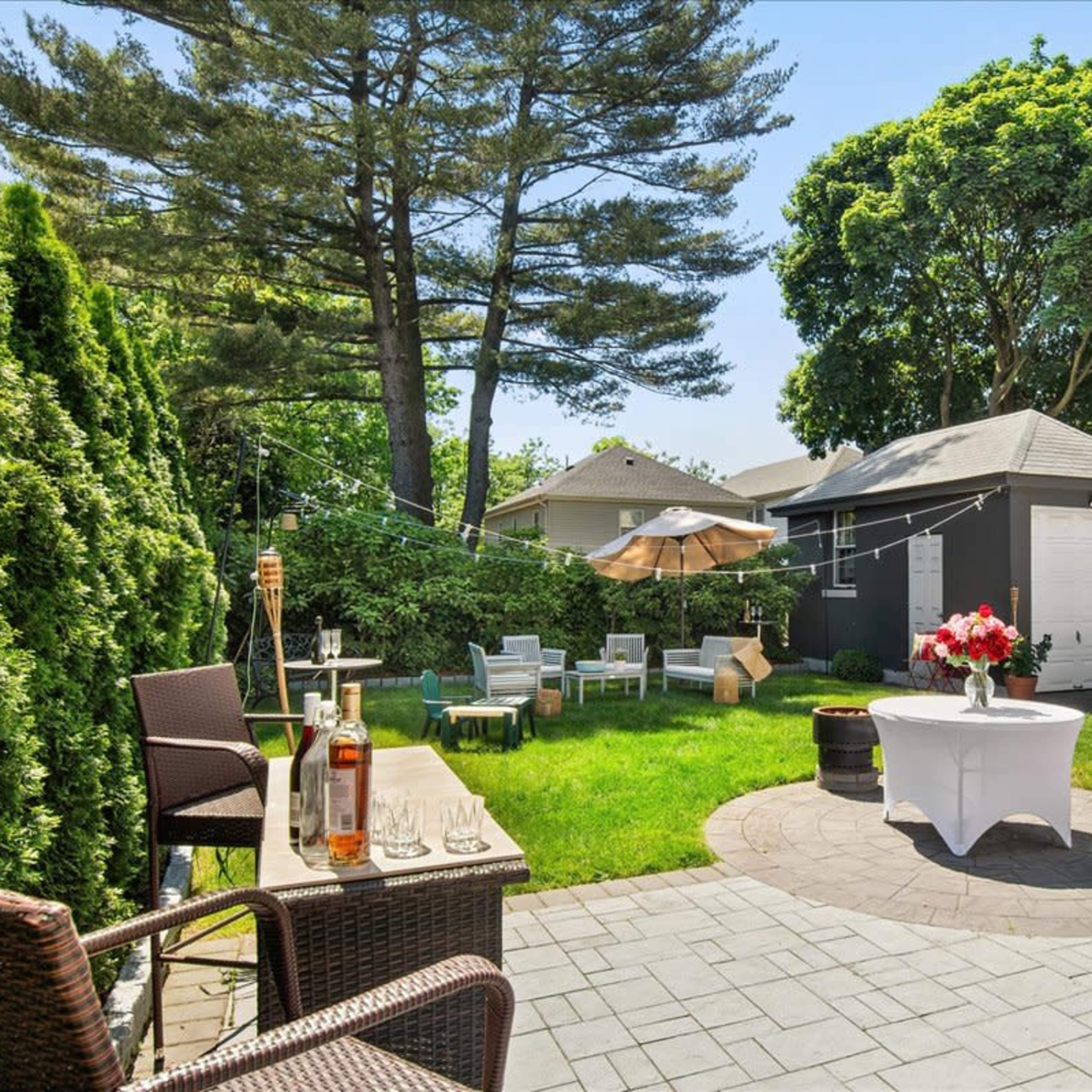 The scene features a landscaped backyard with a stone patio, a green lawn, outdoor seating, a bar area, and umbrellas providing shade.