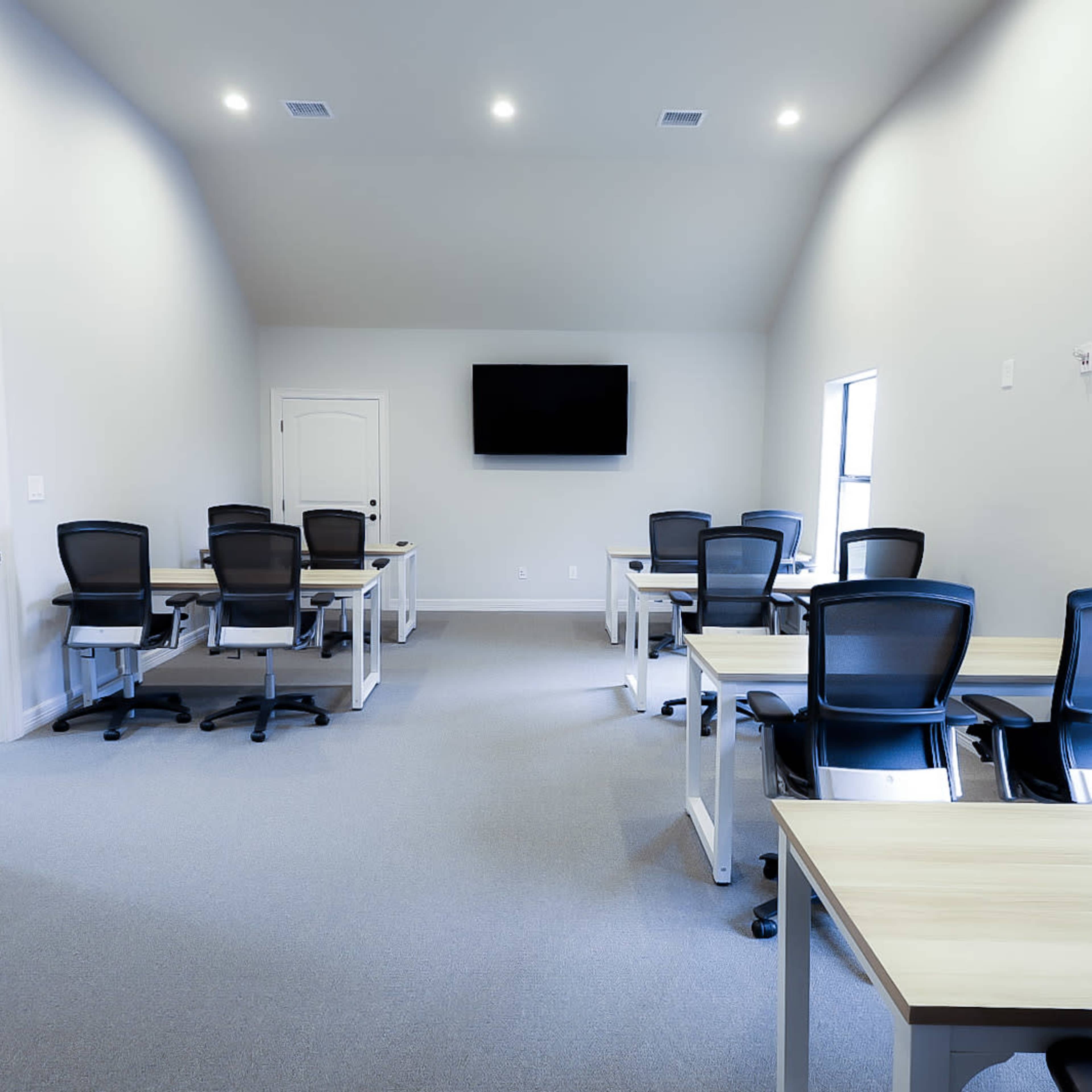 A sparsely furnished meeting room with several desks and chairs arranged in a symmetrical layout, a wall-mounted TV, and windows allowing natural light.