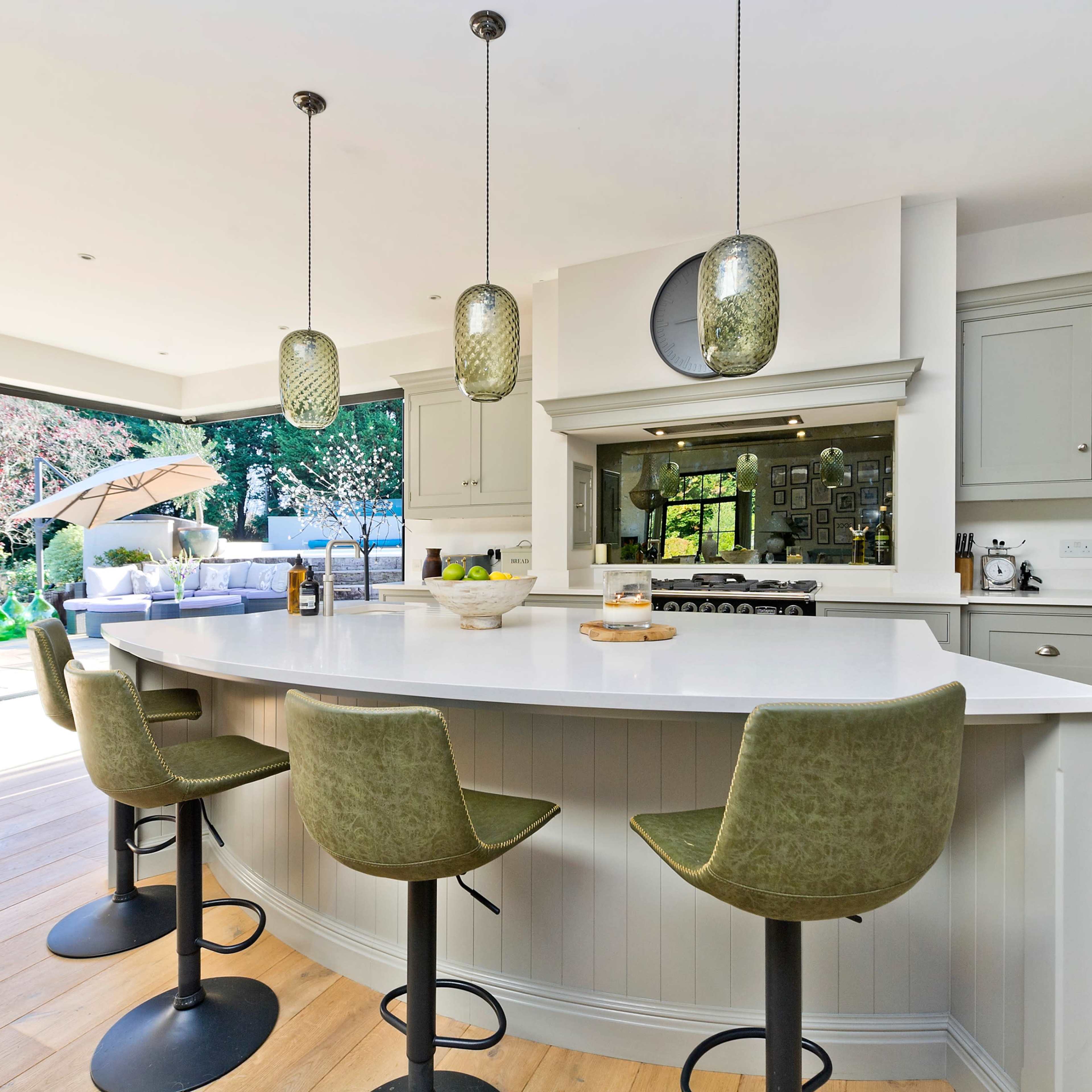 A modern kitchen features a large white island with green bar stools, pendant lighting, and an open view of a garden through glass sliding doors.