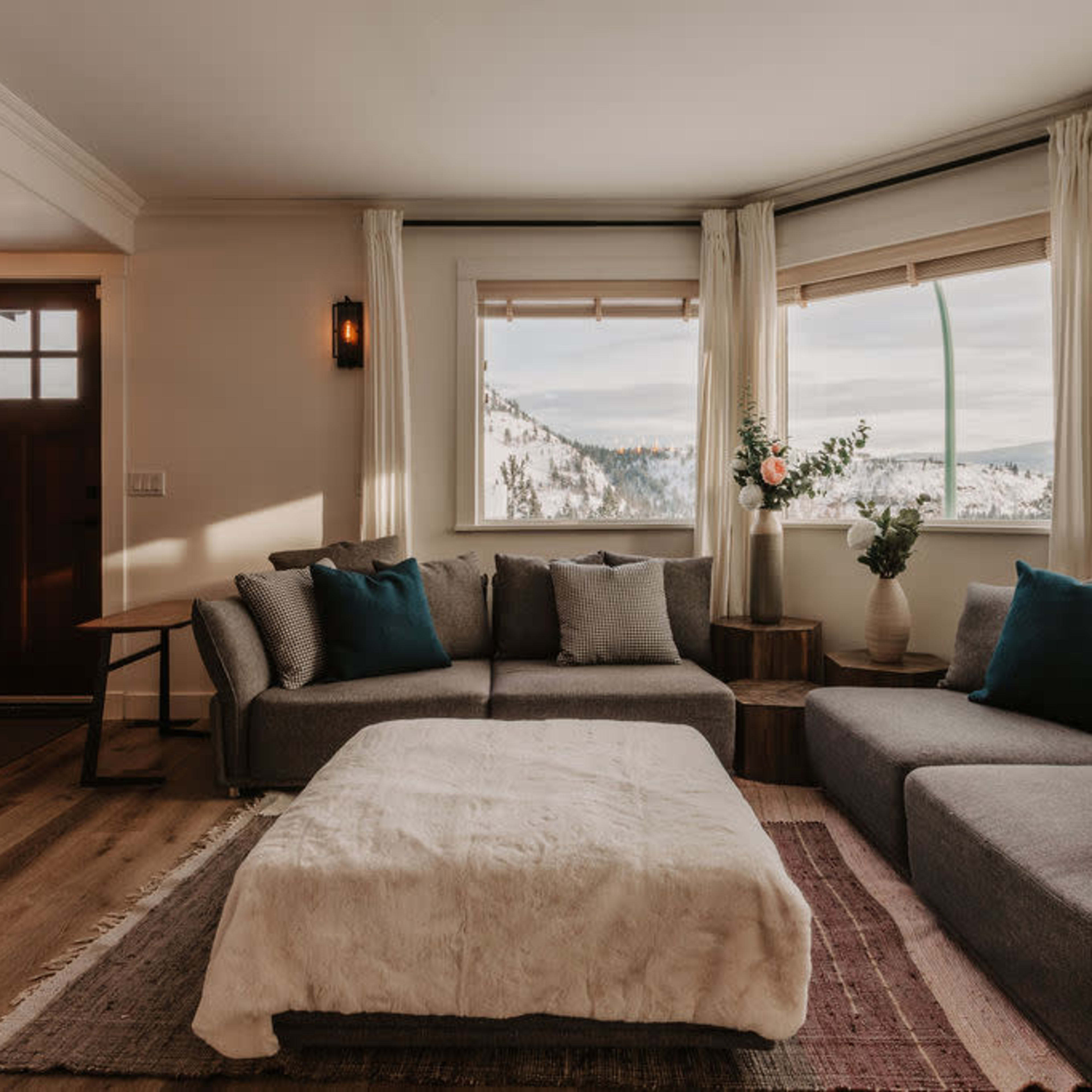 A modern living room features a gray sectional sofa with blue and gray cushions, a light-colored area rug, and large windows showcasing a snowy mountain view.