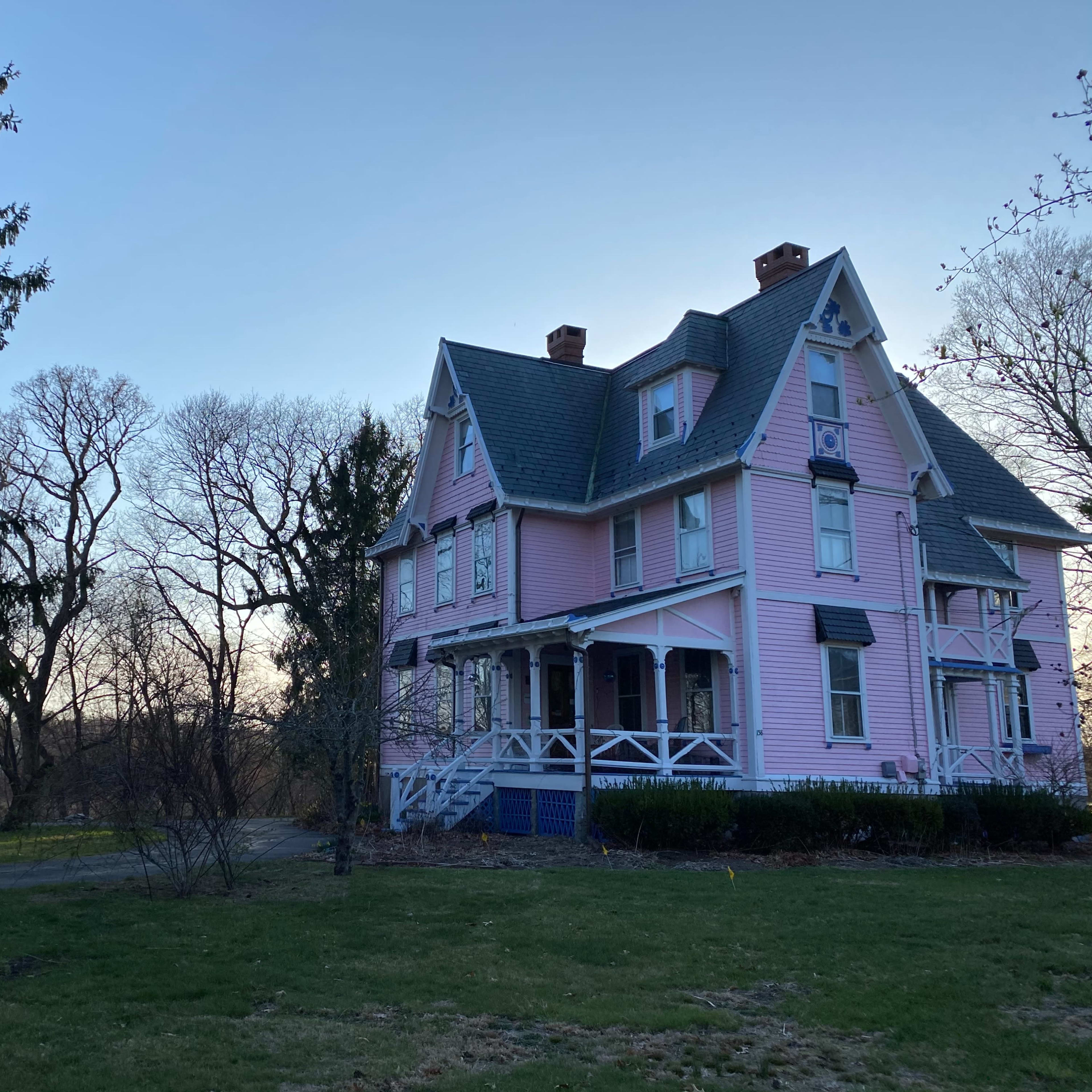 A large pink Victorian house stands at the edge of a grassy area, surrounded by bare trees and a clear sky.