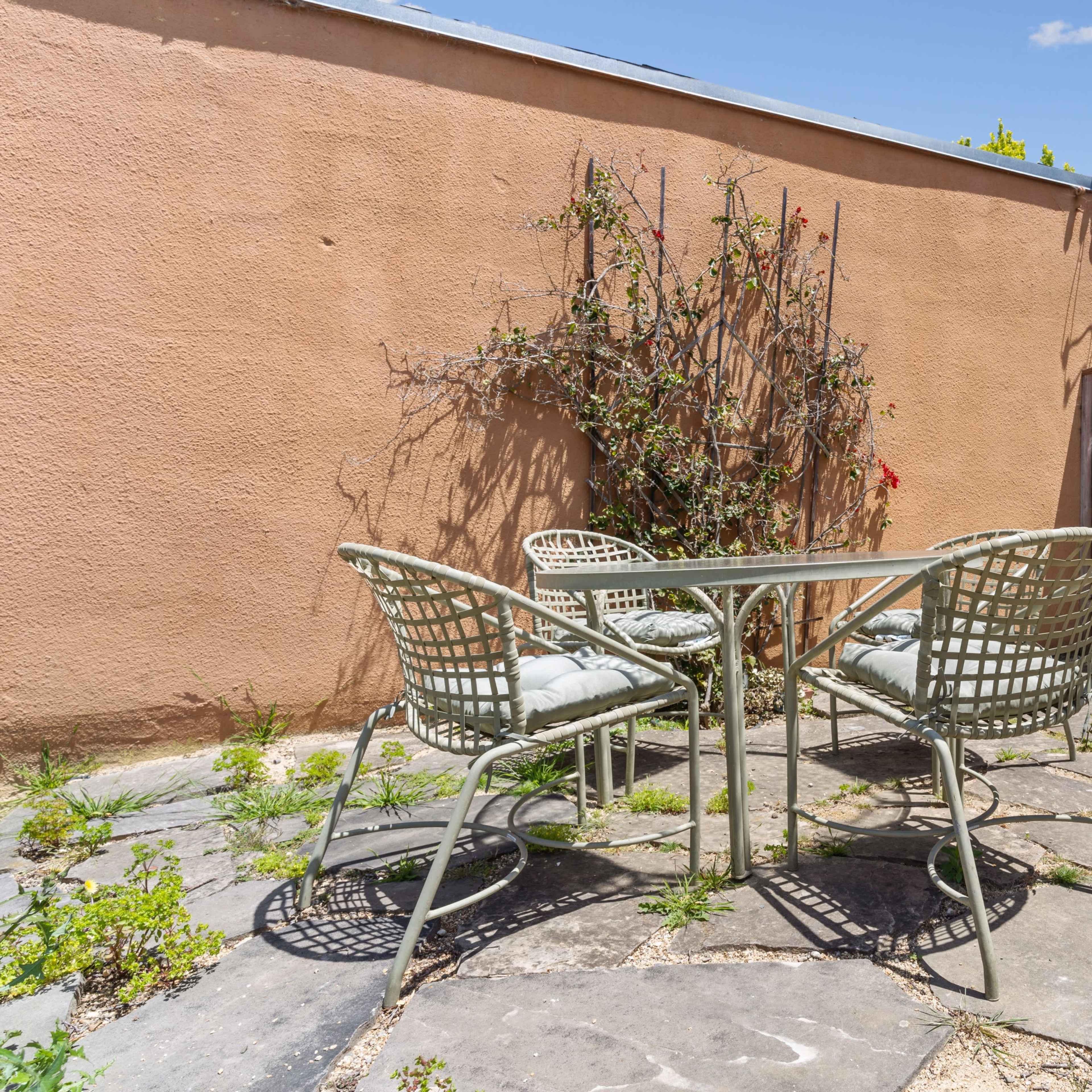 A small outdoor seating area with four wicker chairs and a table is situated on a stone patio surrounded by a wall and a gate.