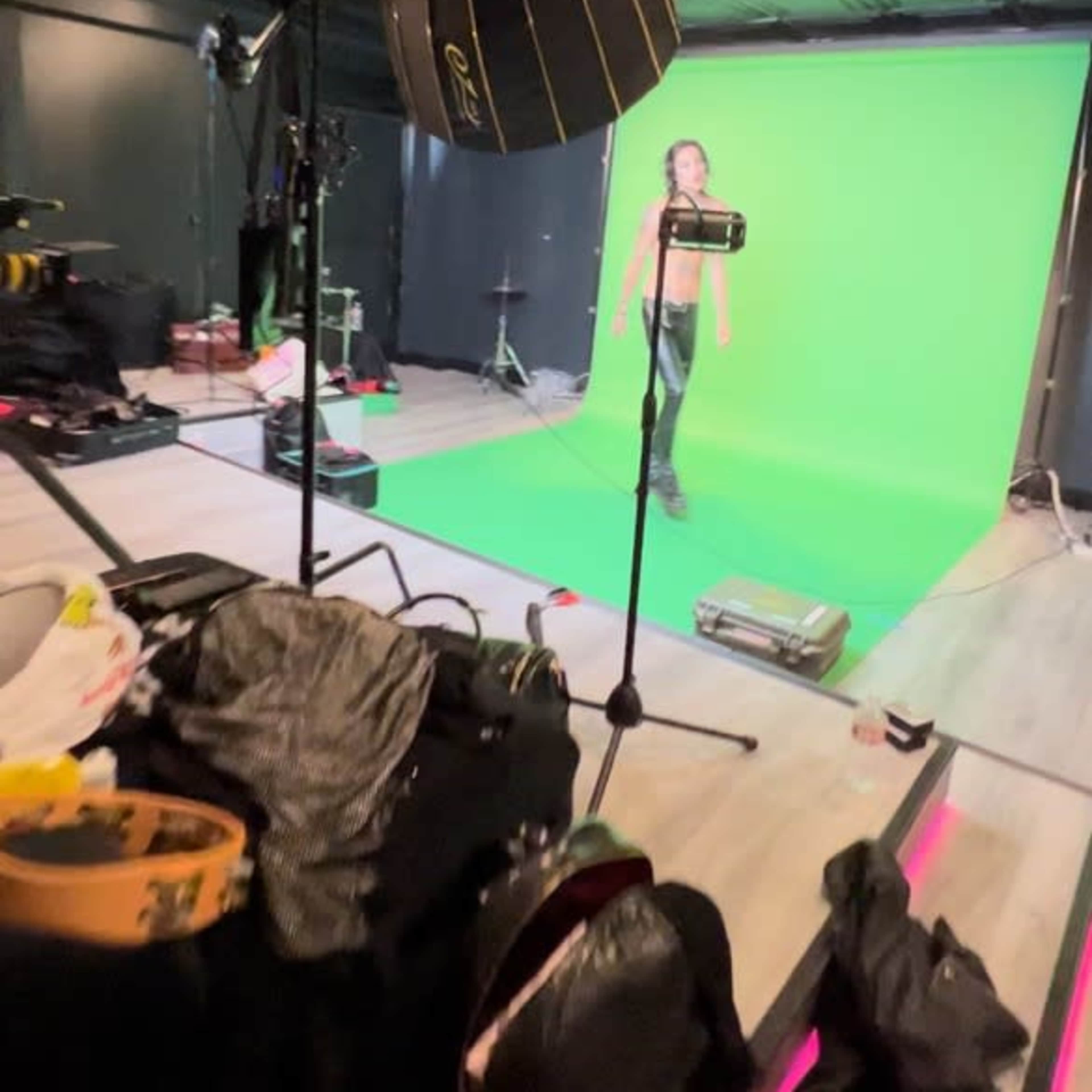 A person stands in front of a green screen in a dimly lit studio, surrounded by camera equipment and various bags on the floor.