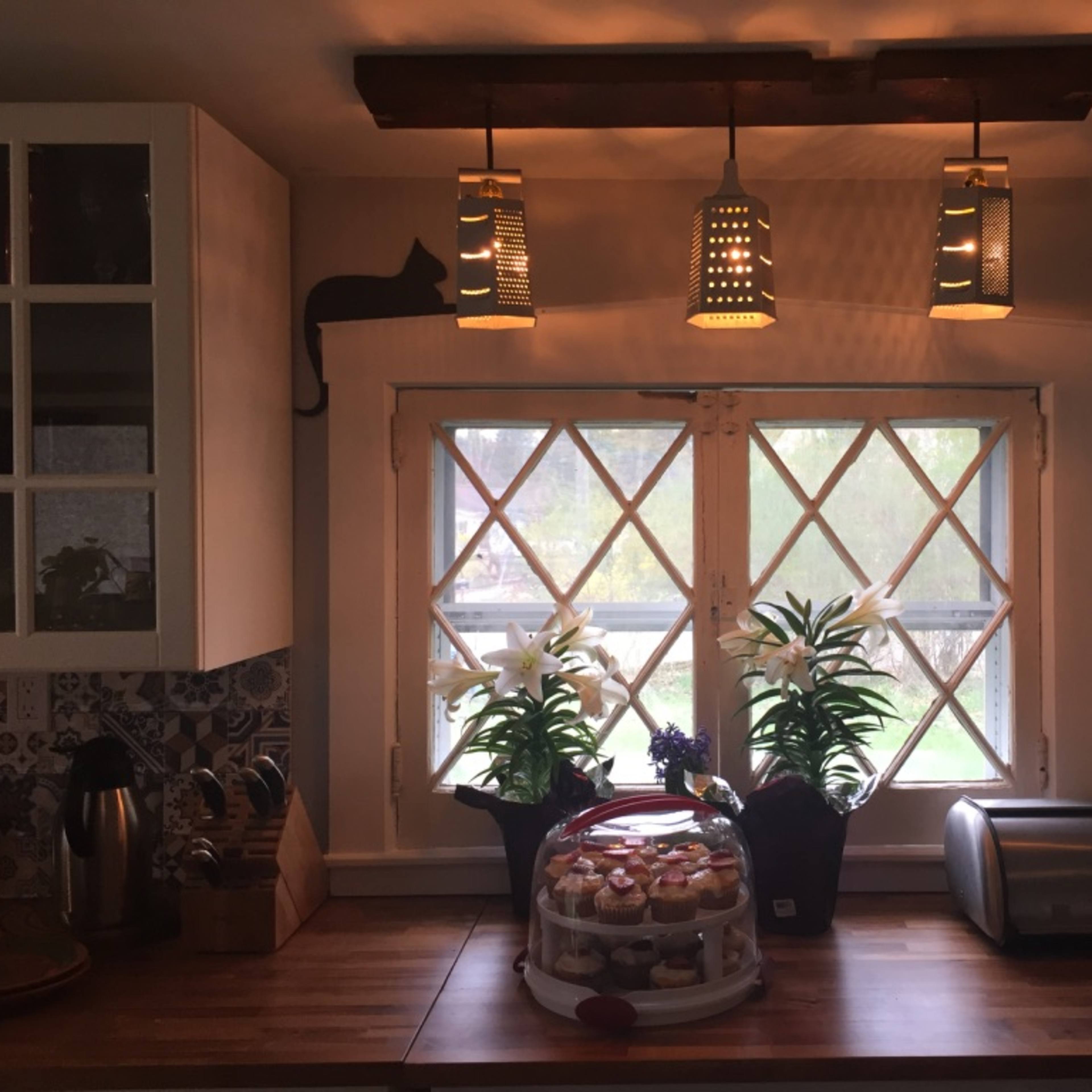 A cozy kitchen features a window adorned with potted lilies and a display of cupcakes under a glass dome, illuminated by hanging lanterns.