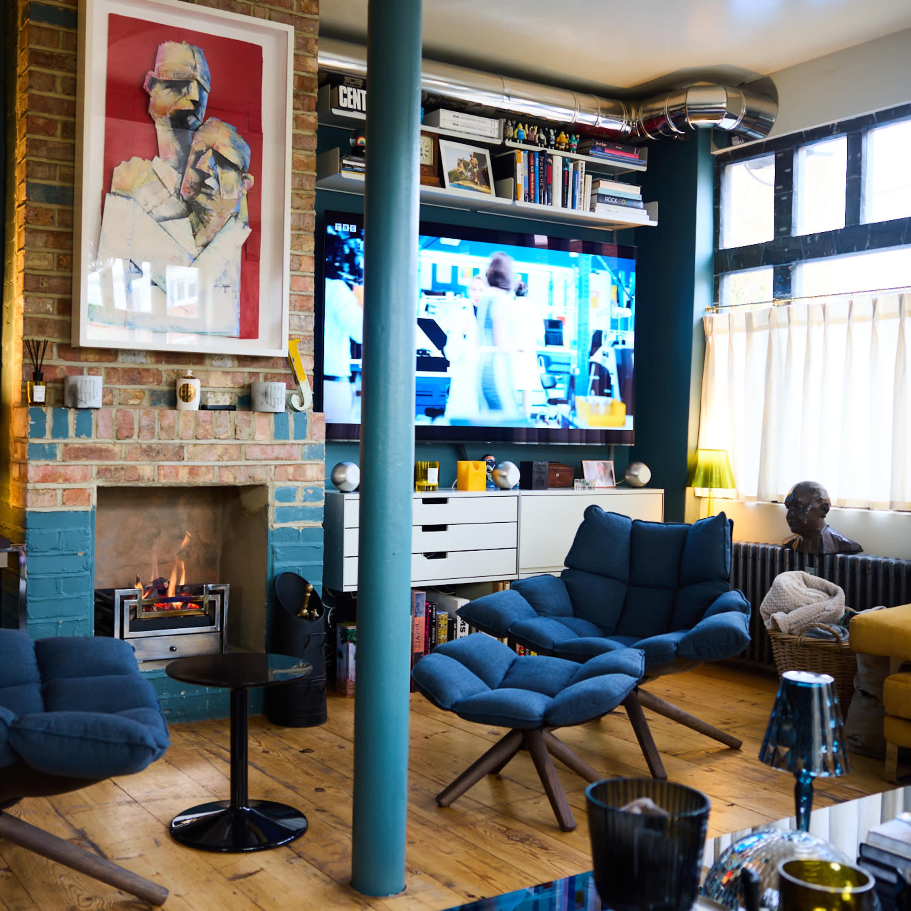 A living room with a brick fireplace, two blue armchairs, a television mounted on the wall, and various artworks and bookshelves.