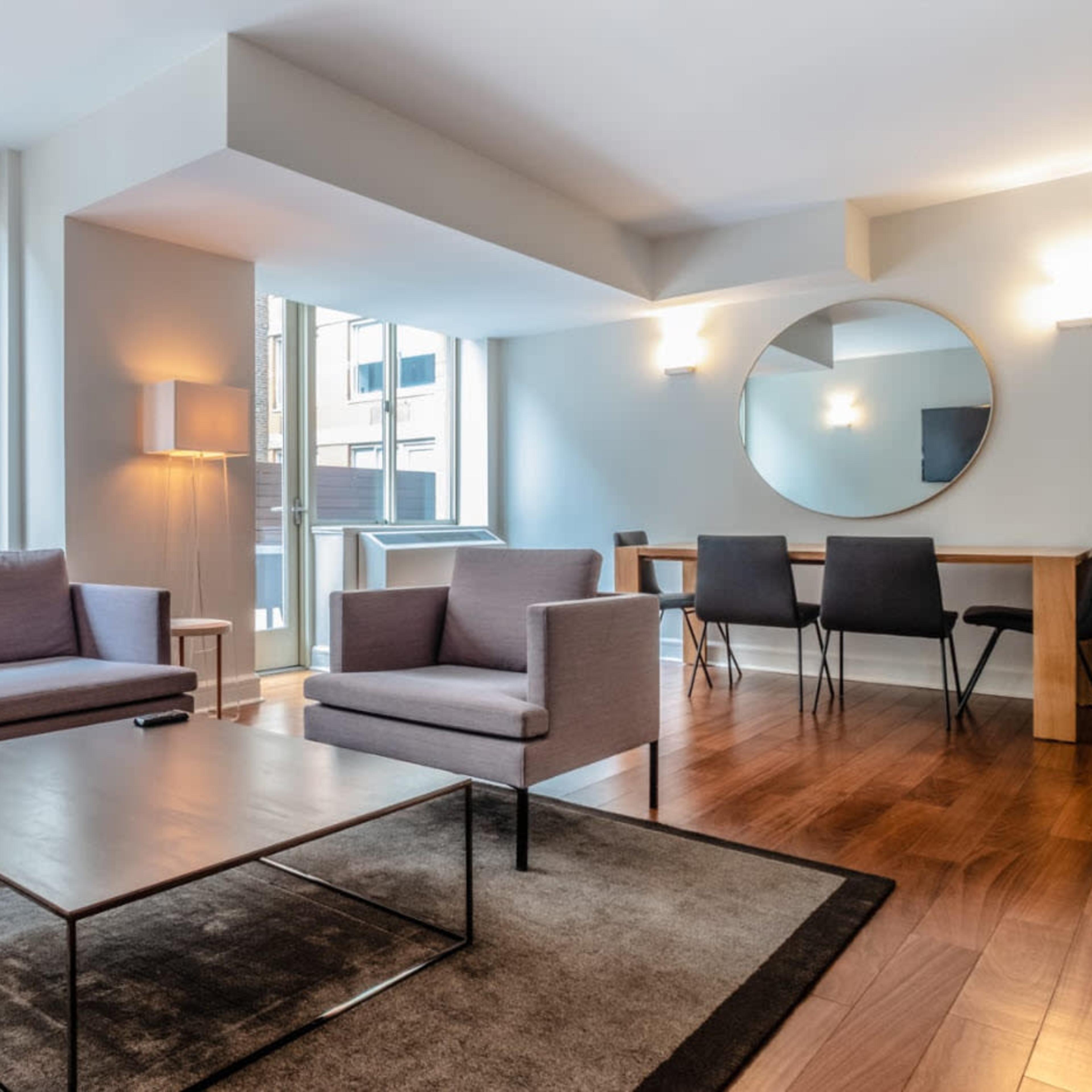 A modern living area with a gray sofa, chairs, a glass coffee table, and a dining table in a well-lit space with large windows.