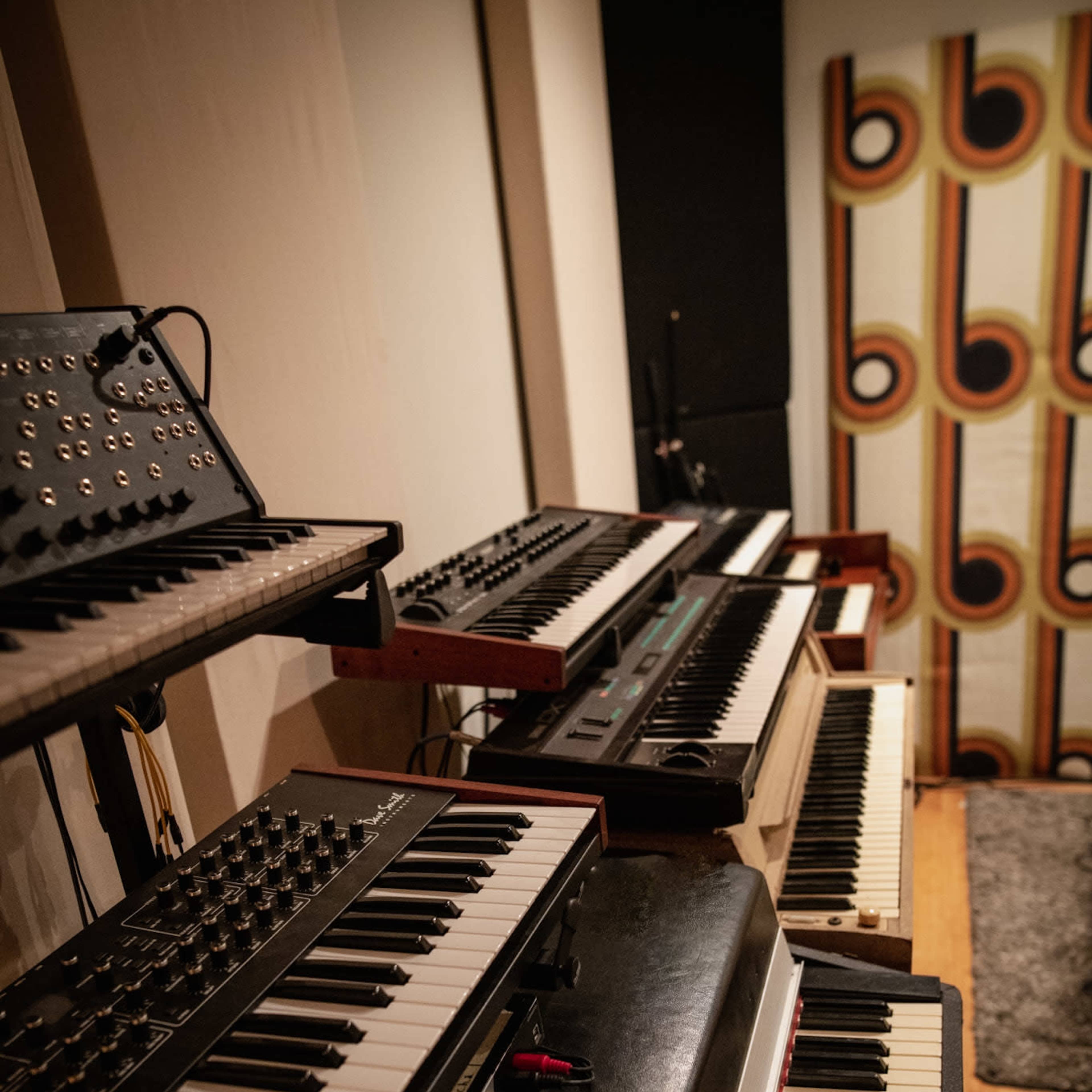 A collection of synthesizers and keyboards arranged in a music studio, with a patterned backdrop and wooden flooring.