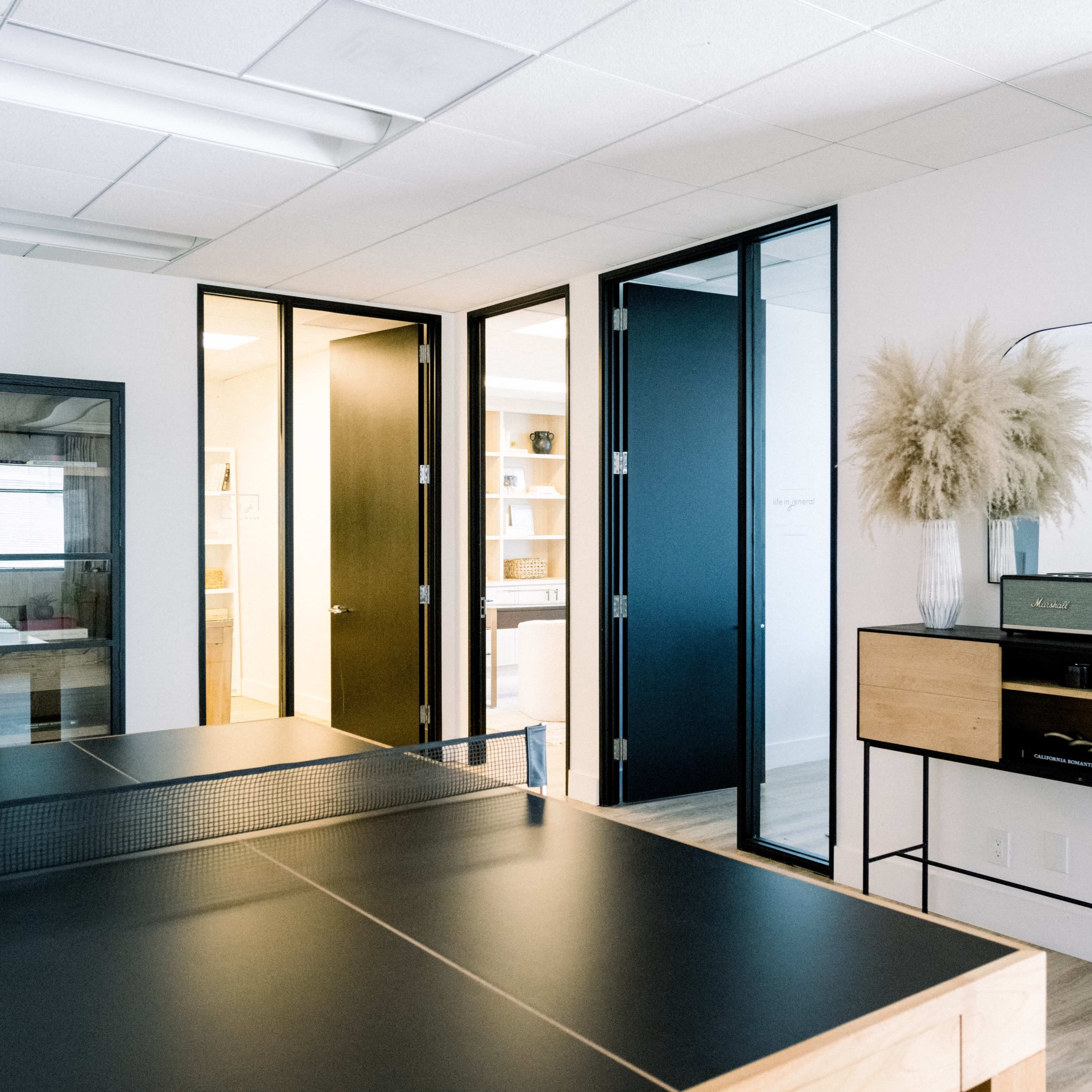 A modern office space features a ping pong table, black and wood furniture, and several closed doors leading to other areas.
