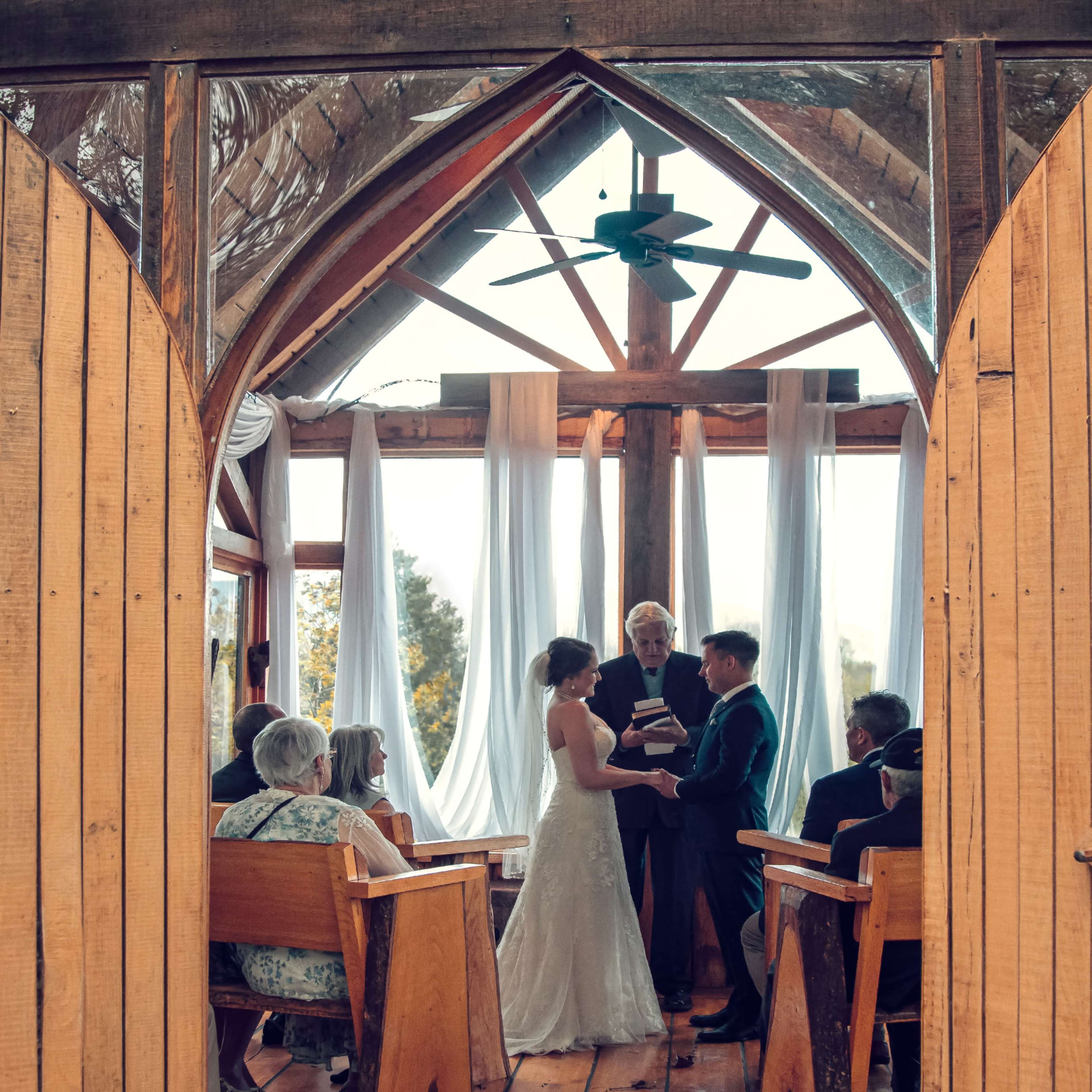 A couple stands at the altar inside a wooden chapel, surrounded by guests, as they exchange vows under a large ceiling fan.