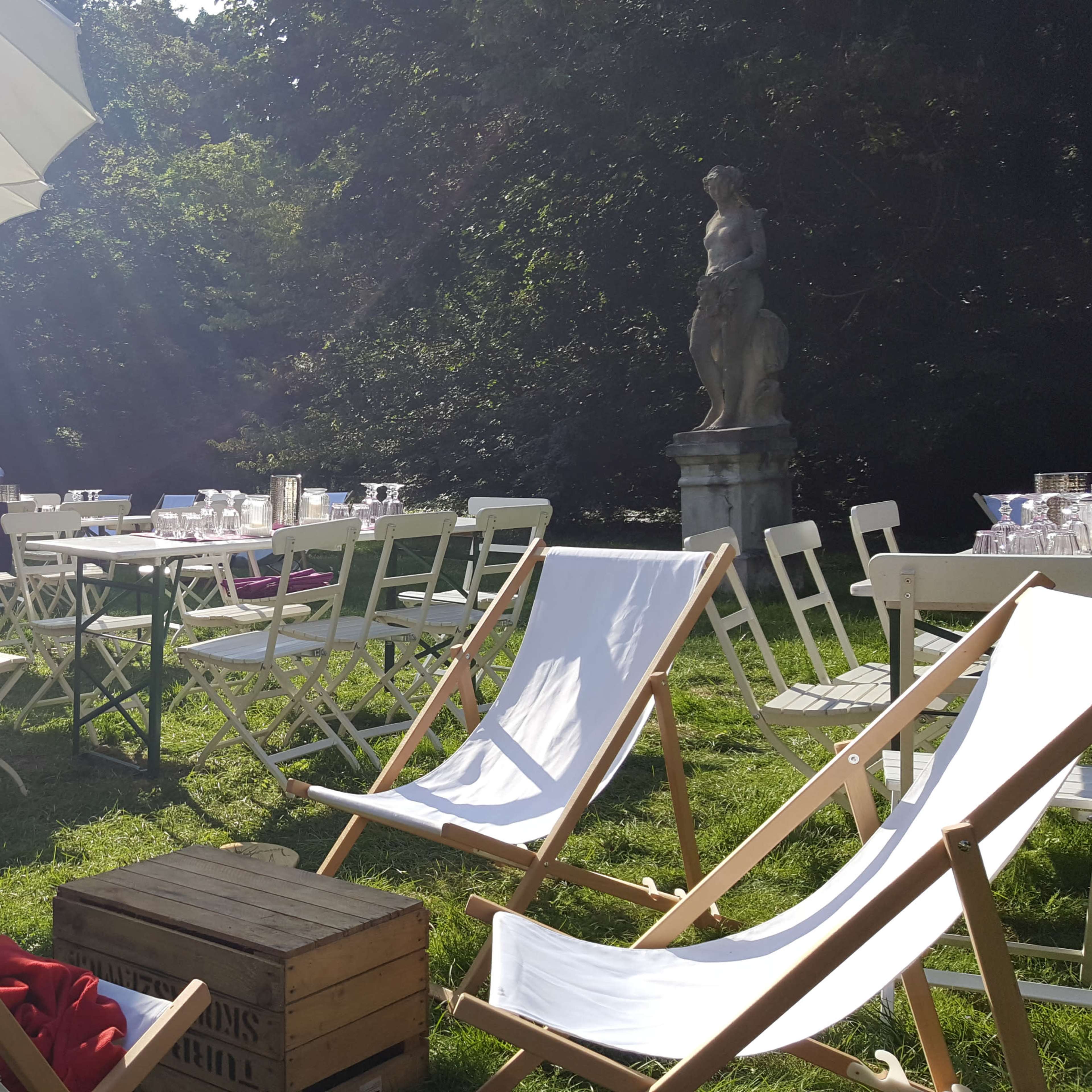 The scene depicts an outdoor event area featuring rows of white tables and chairs, a large umbrella, and lounge chairs arranged on green grass.