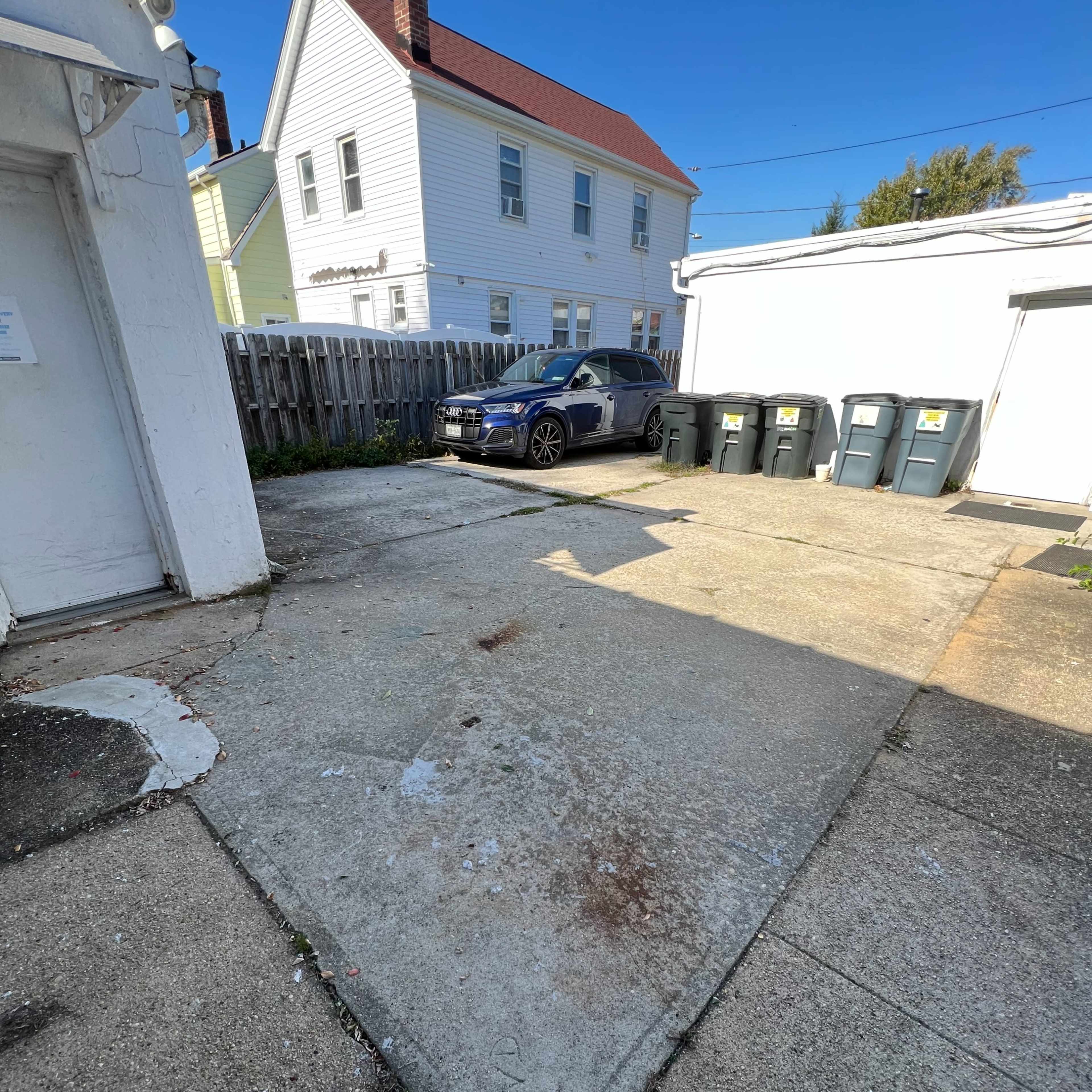 A concrete parking area next to a white house with a red roof and several gray trash bins lined up against a garage.
