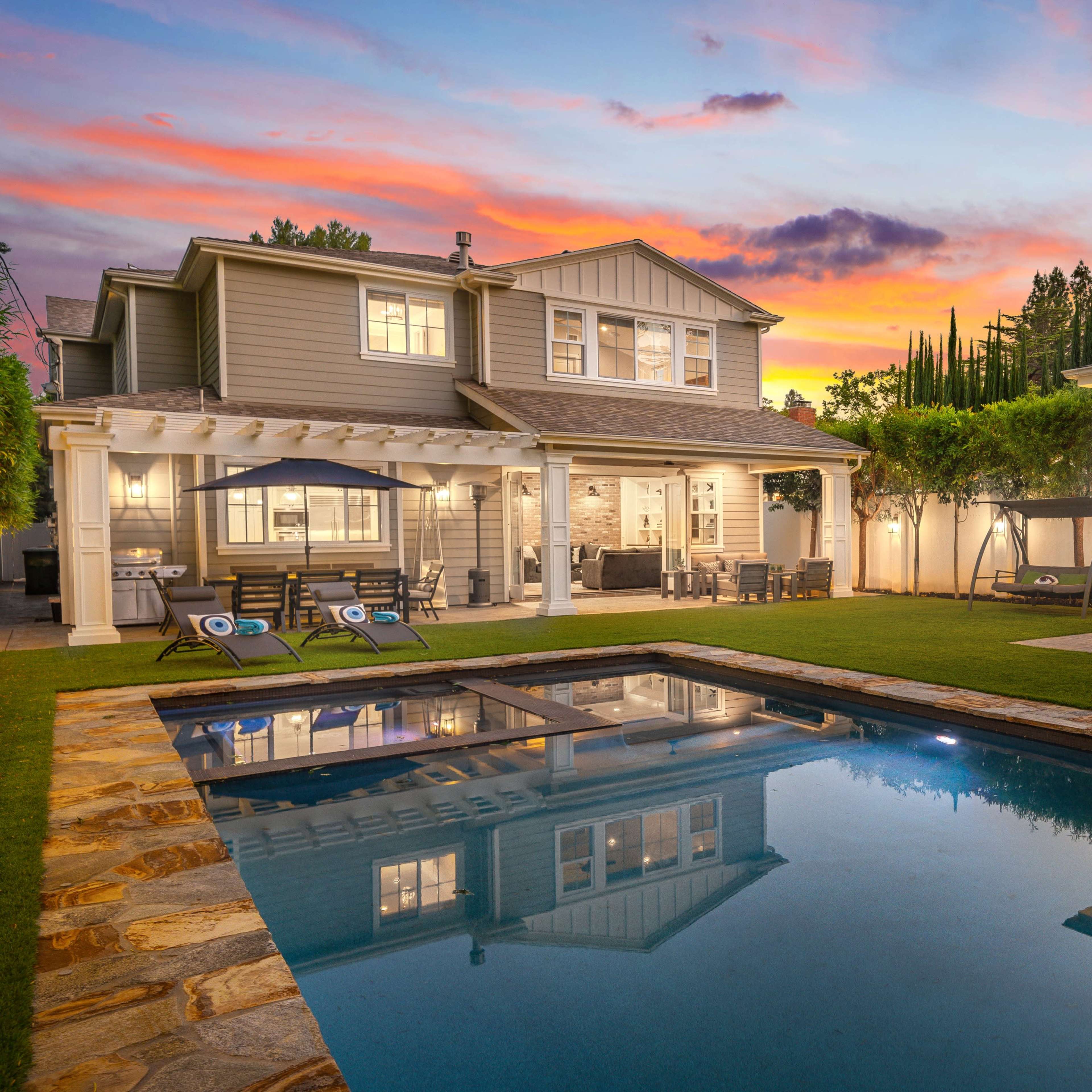 A modern two-story house with a landscaped backyard featuring a swimming pool and a colorful sunset sky.