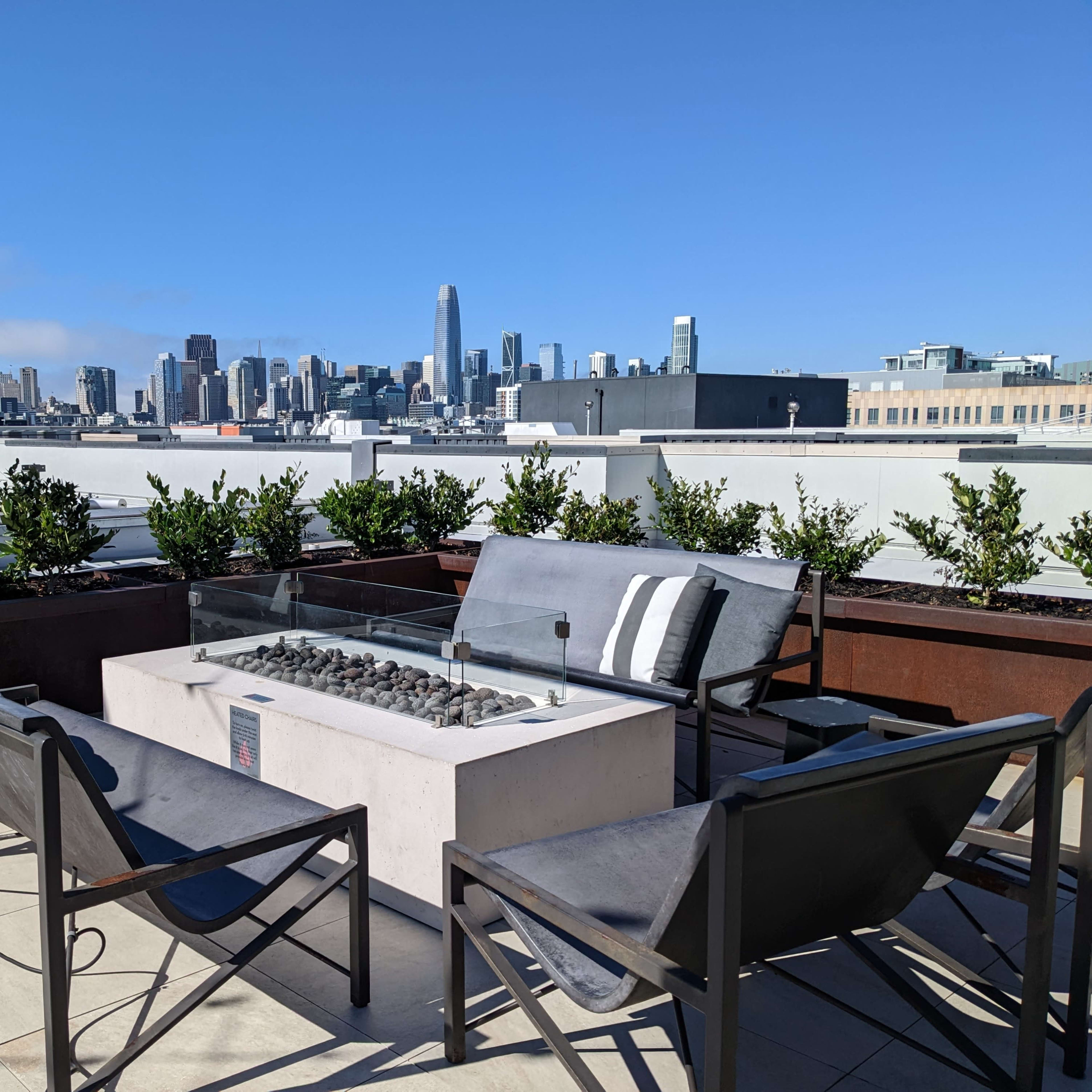 A rooftop patio features a fire pit surrounded by chairs and a view of a city skyline.