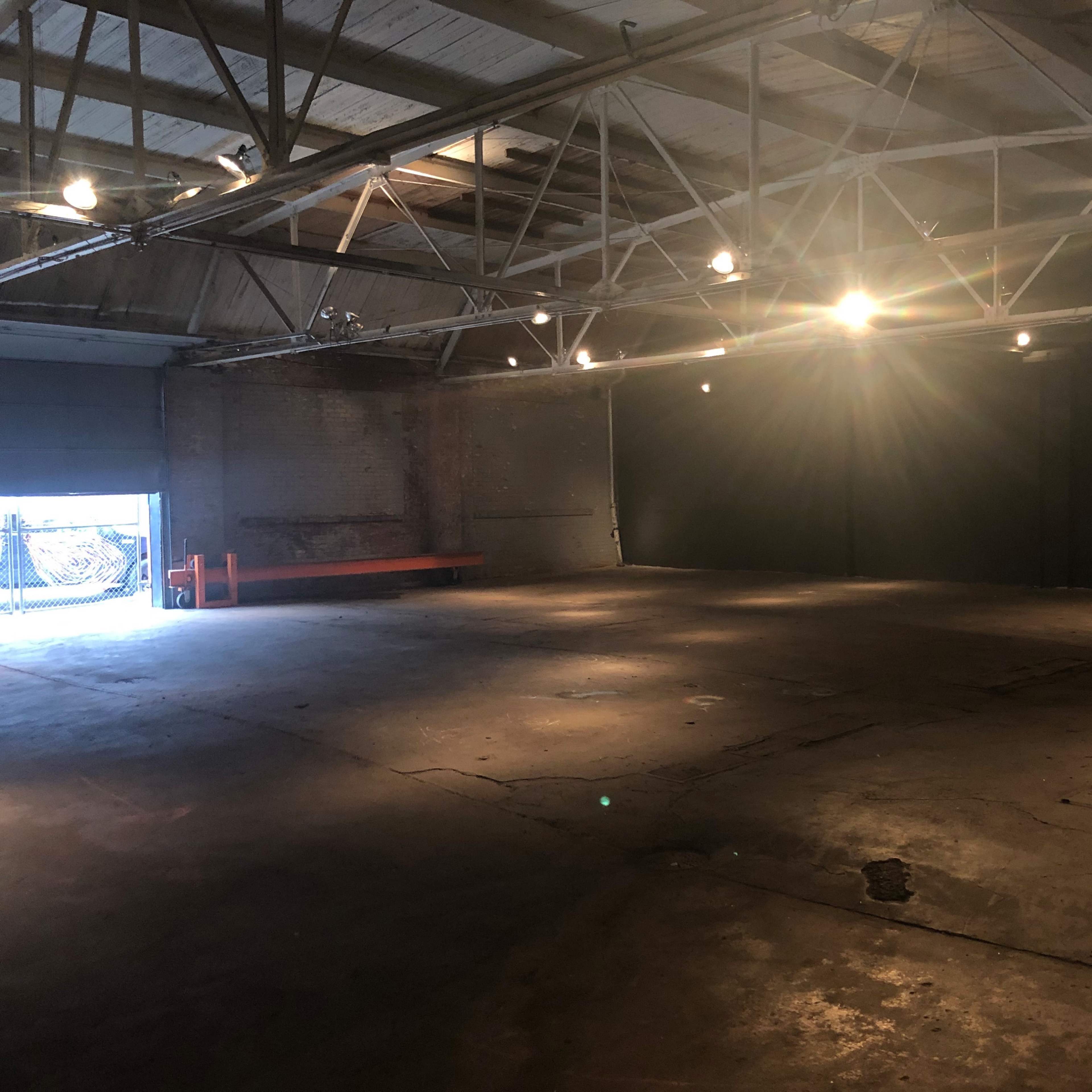 An empty industrial space with a concrete floor, exposed beams, and lighting fixtures, featuring a doorway that leads to an exterior area.