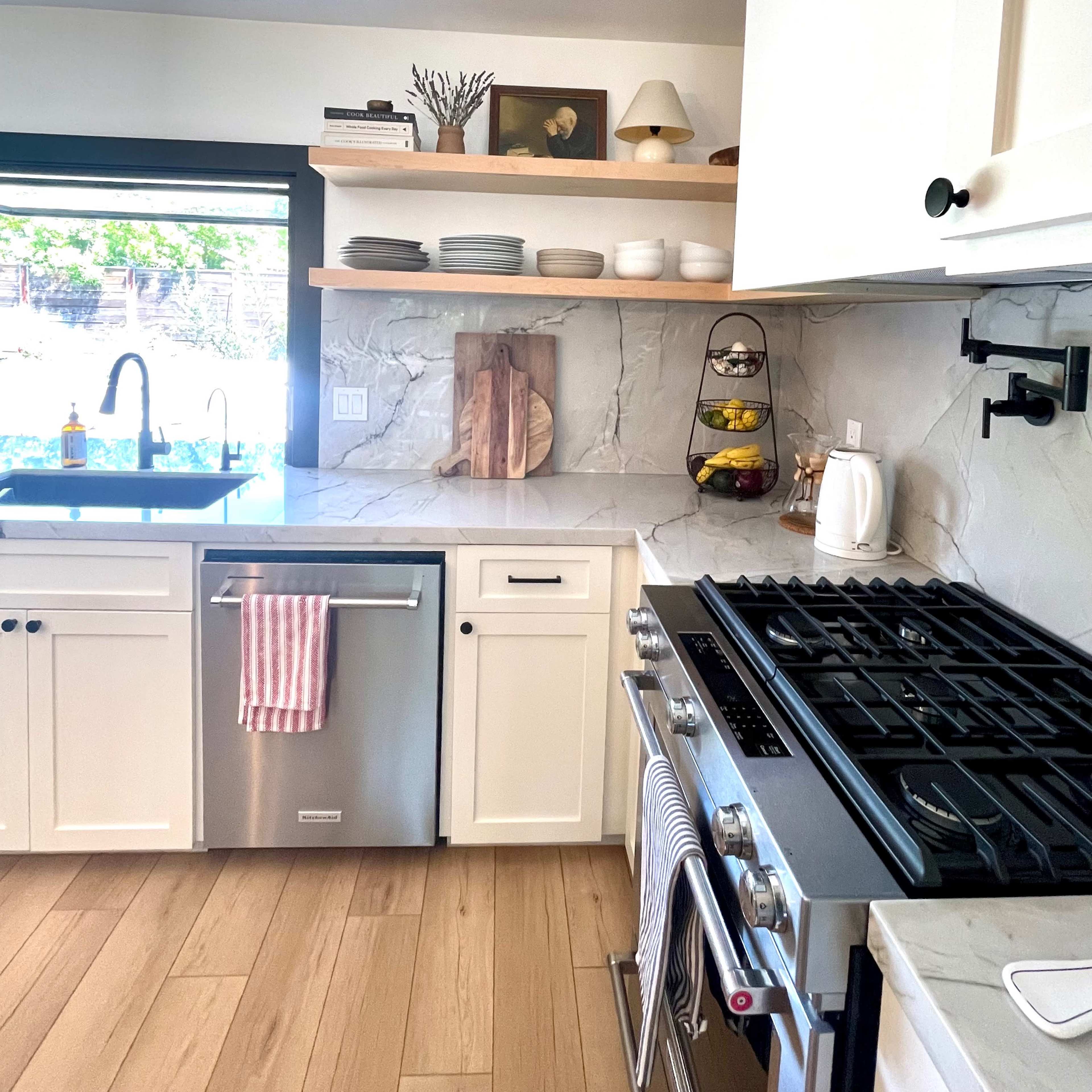 A modern kitchen with white cabinetry, a stainless steel dishwasher, and a gas stove, featuring open shelving above a counter with decorative items.