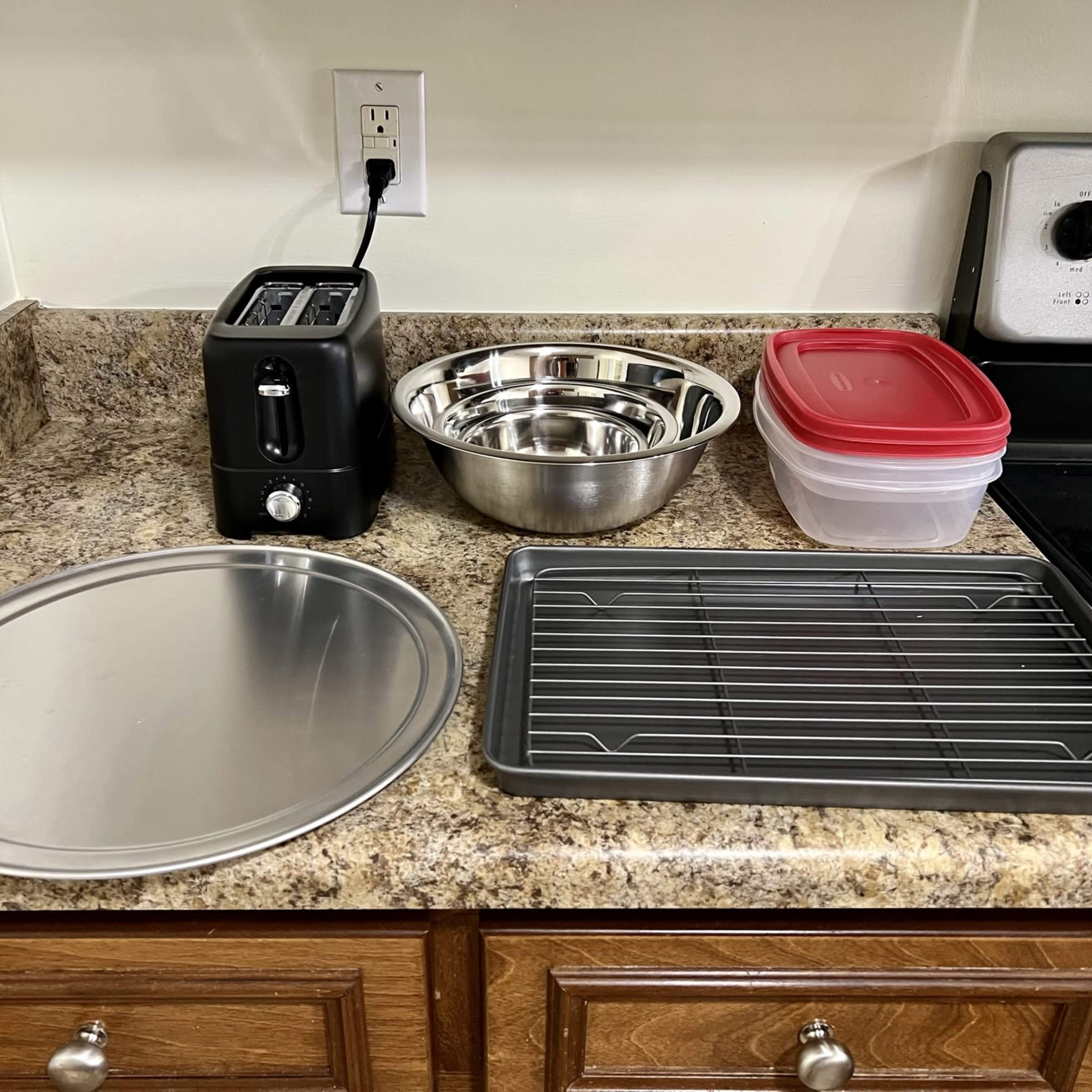 A kitchen countertop features a toaster, several metal bowls, a round baking pan, a cooling rack, and a plastic container with a red lid.
