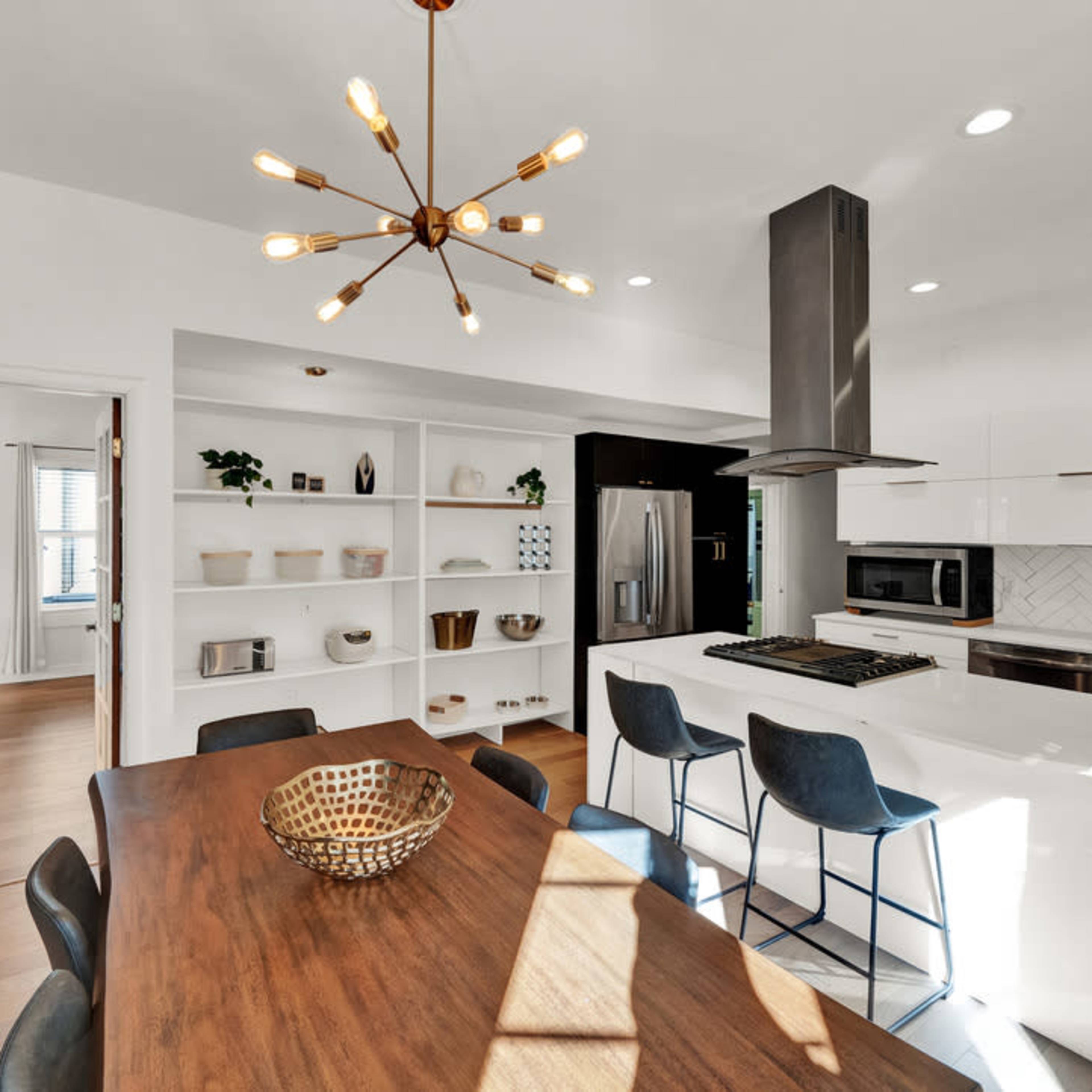 A modern kitchen with white cabinetry, a stainless steel hood, and a dining table set with a decorative bowl, illuminated by pendant lights.