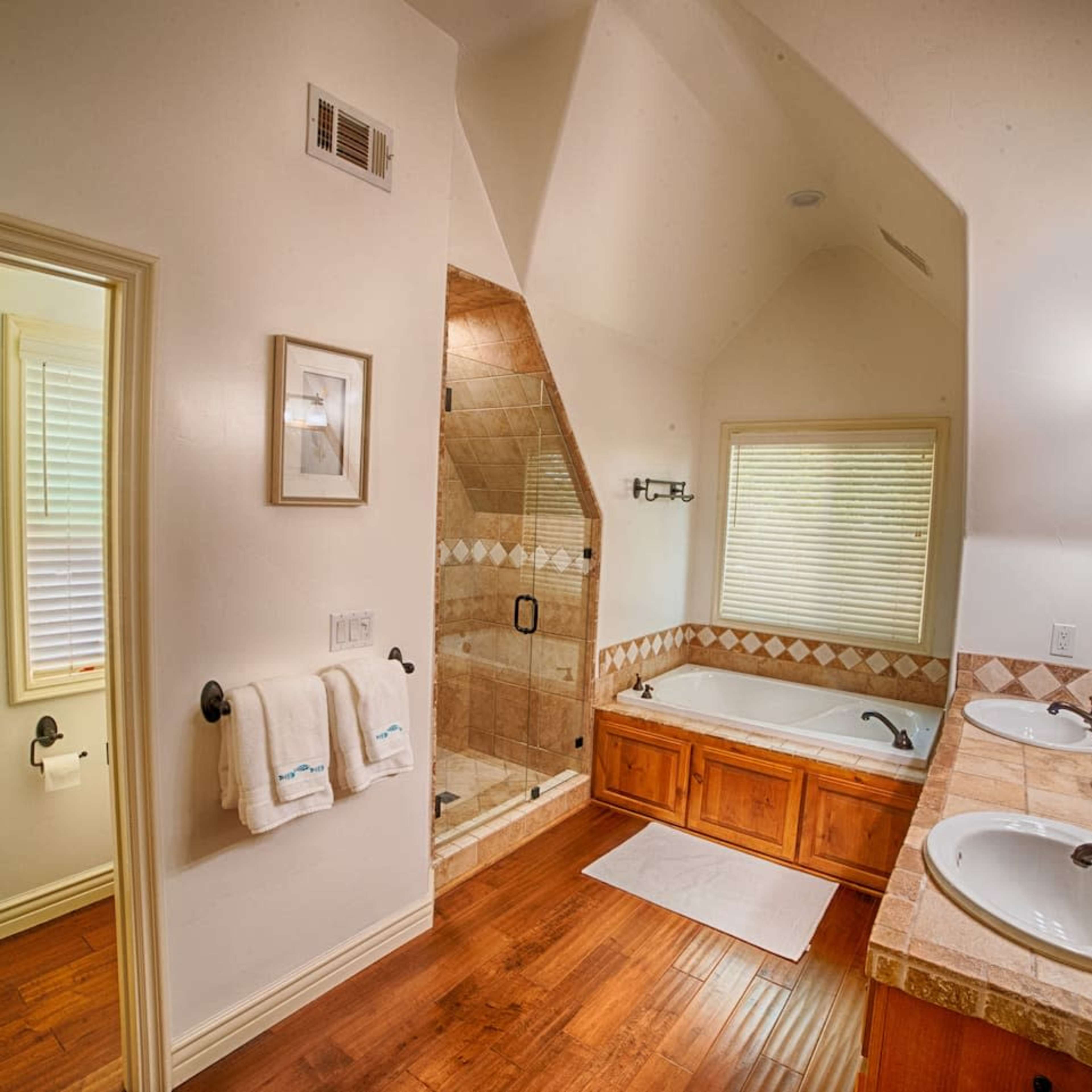 A bathroom featuring a double vanity with sinks, a corner bathtub, and a separate shower area, all complemented by wooden flooring and natural light from windows.