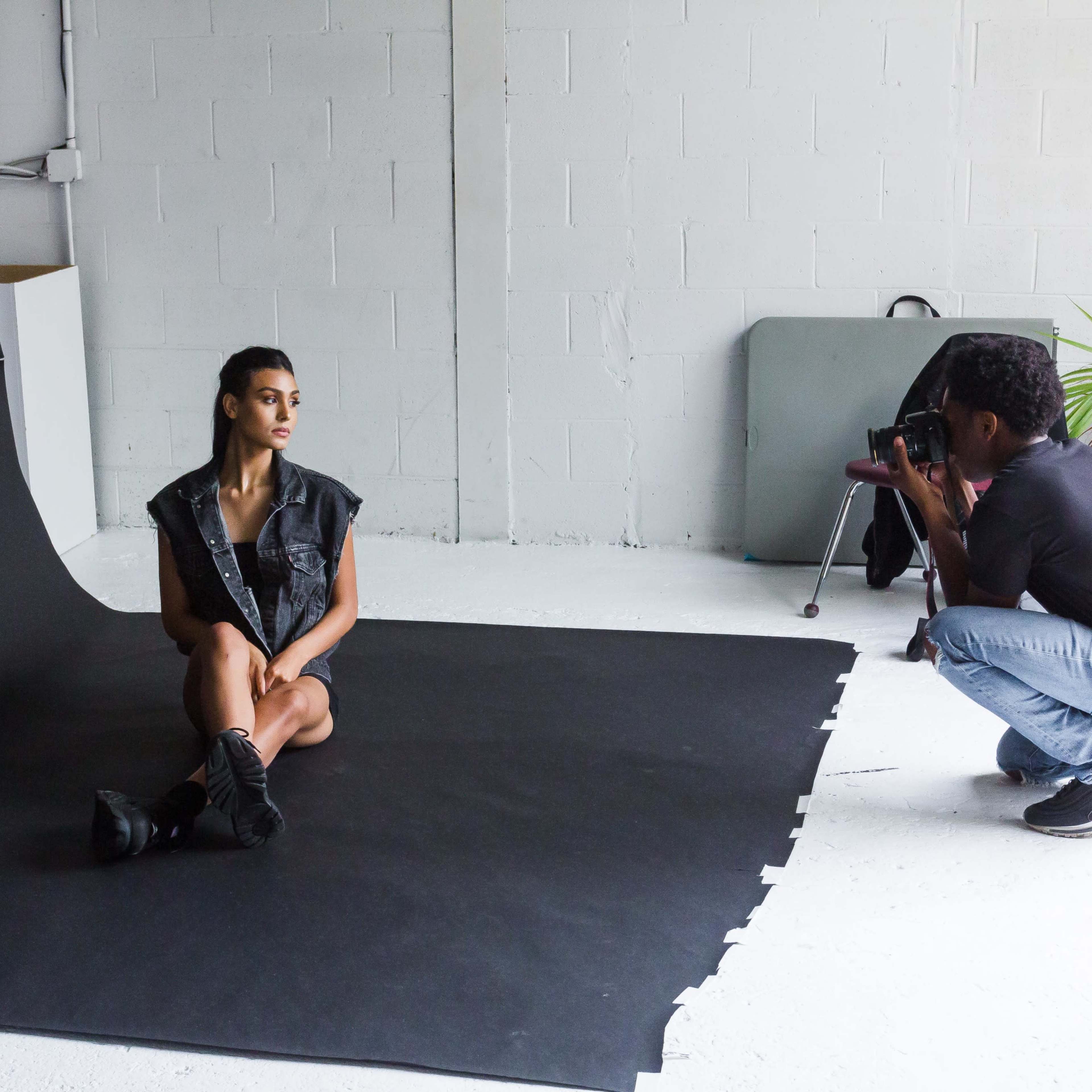 A photographer is taking pictures of a model seated on a black backdrop in a studio with greenery and various furniture.