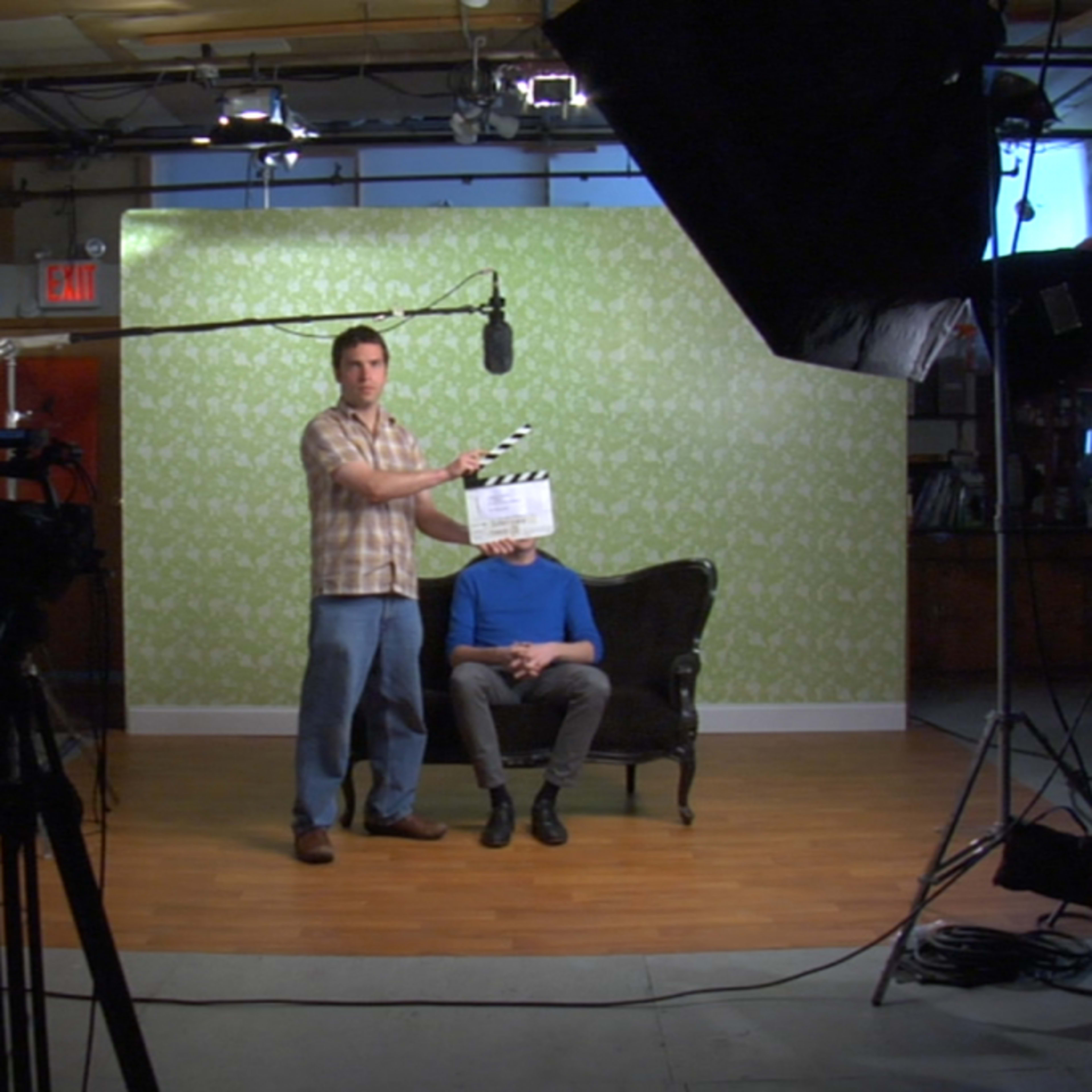A man holds a clapperboard in front of a seated individual on a set with a green patterned background and various lighting equipment.