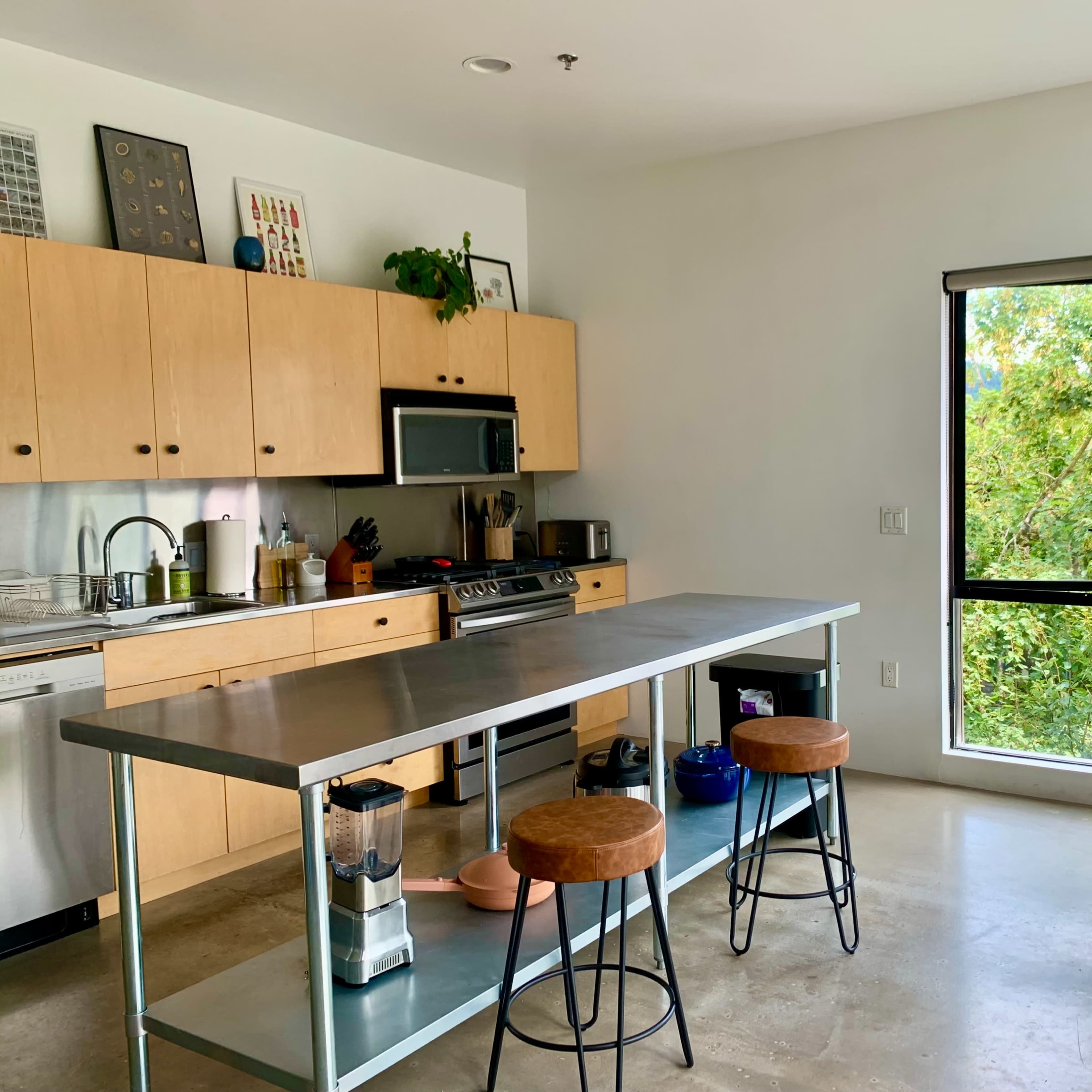 A modern kitchen with wooden cabinets, a stainless steel stove, and a dining area featuring a round table and chairs, with a window overlooking greenery.