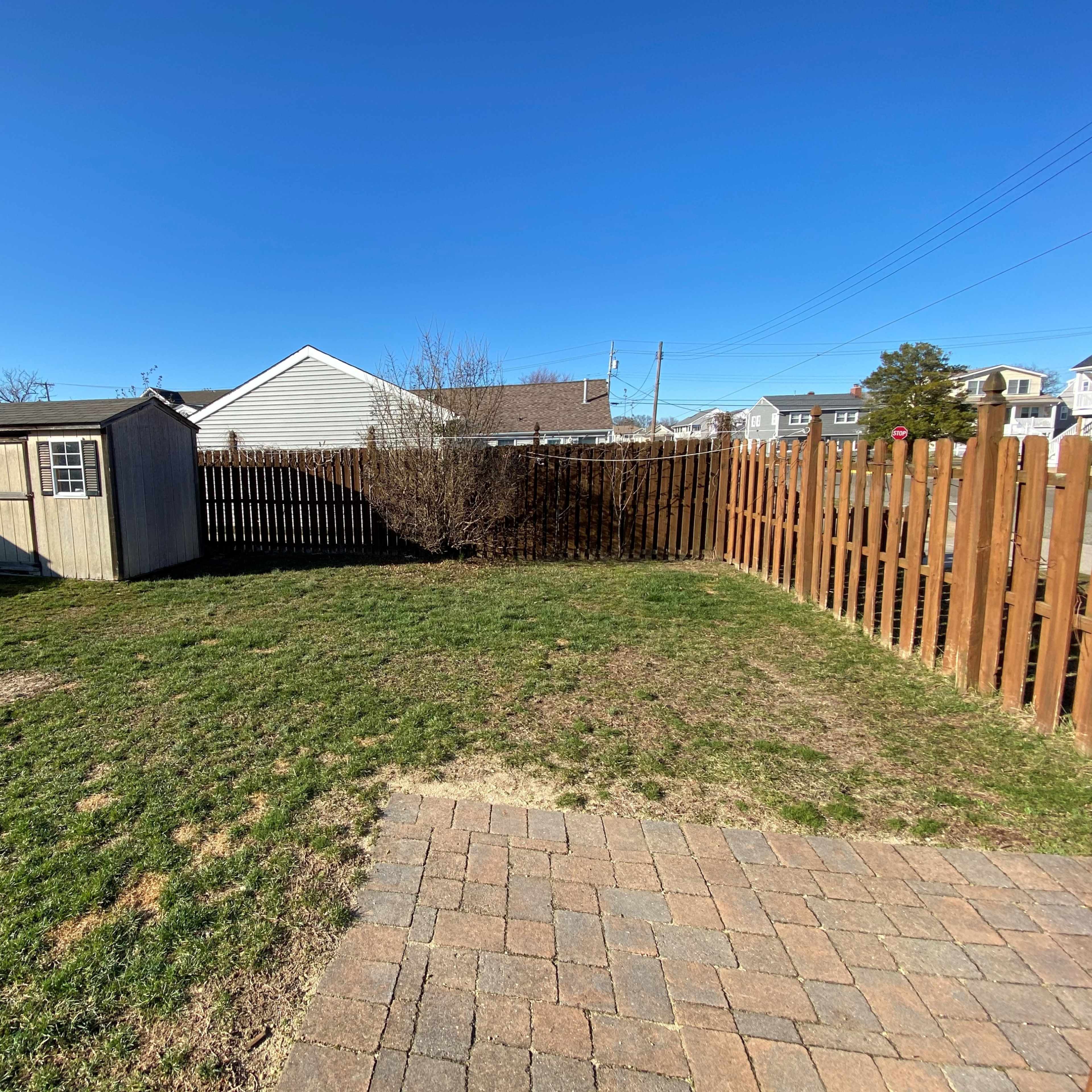A backyard with a small shed, a fence, and a paved area, set against a clear blue sky.