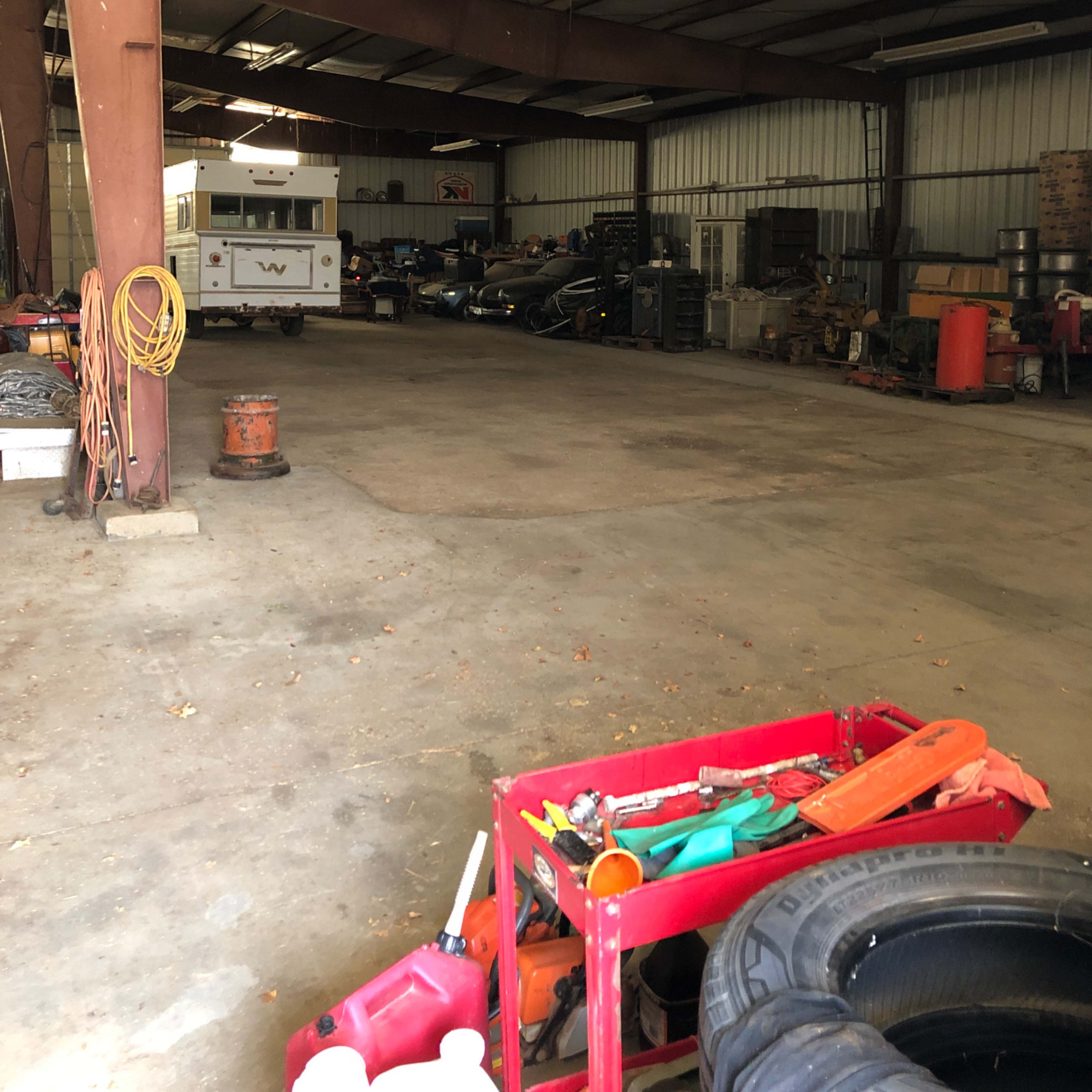 A spacious metal building interior, featuring various tools and equipment scattered throughout, with some vehicles parked along the back.