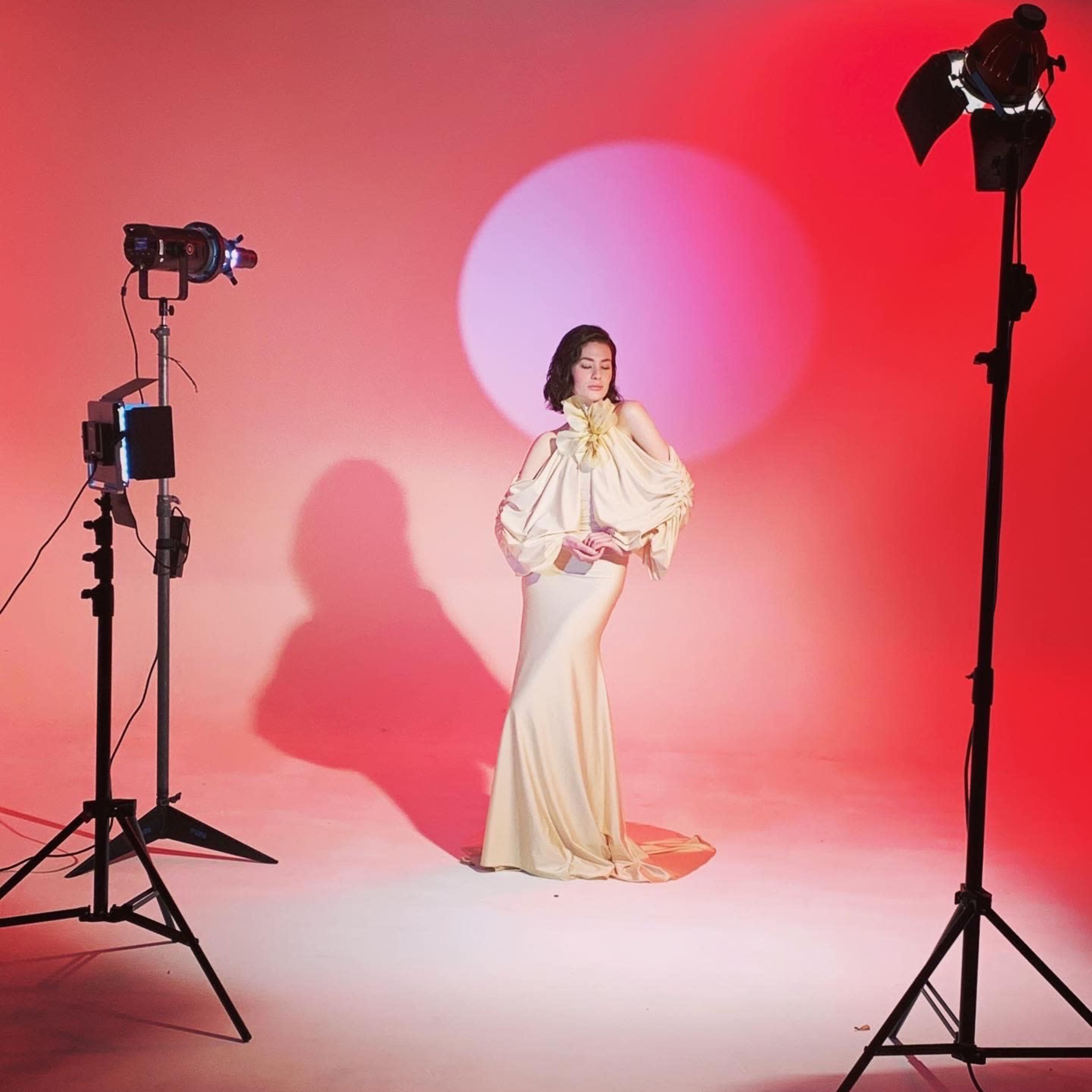 A model poses in an elegant gown against a gradient backdrop while being illuminated by studio lights.