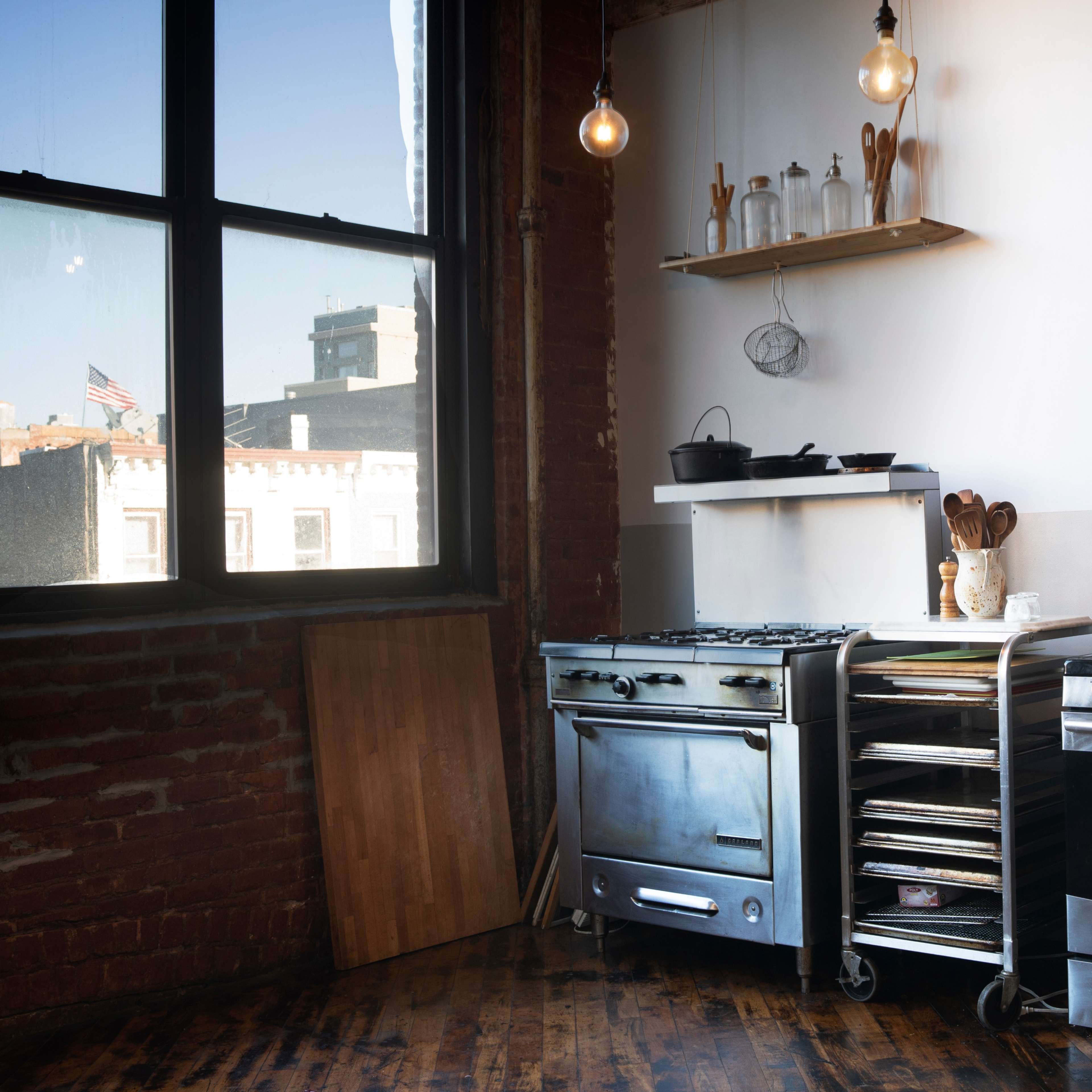 A kitchen with stainless steel appliances, wooden shelves holding kitchenware, and a large window illuminating the space.