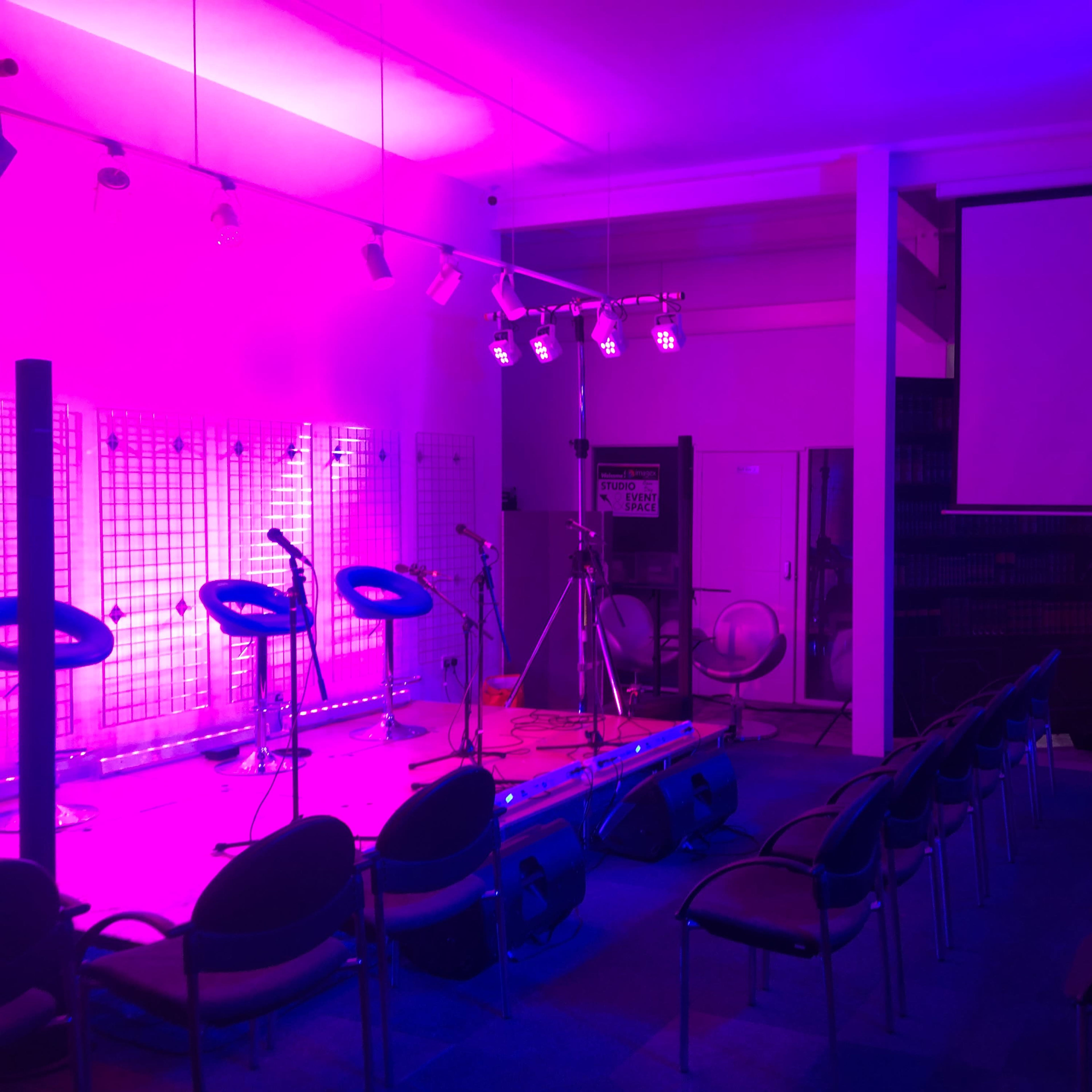 A performance space with purple lighting, featuring a stage with microphones, cameras, and chairs arranged for an audience.