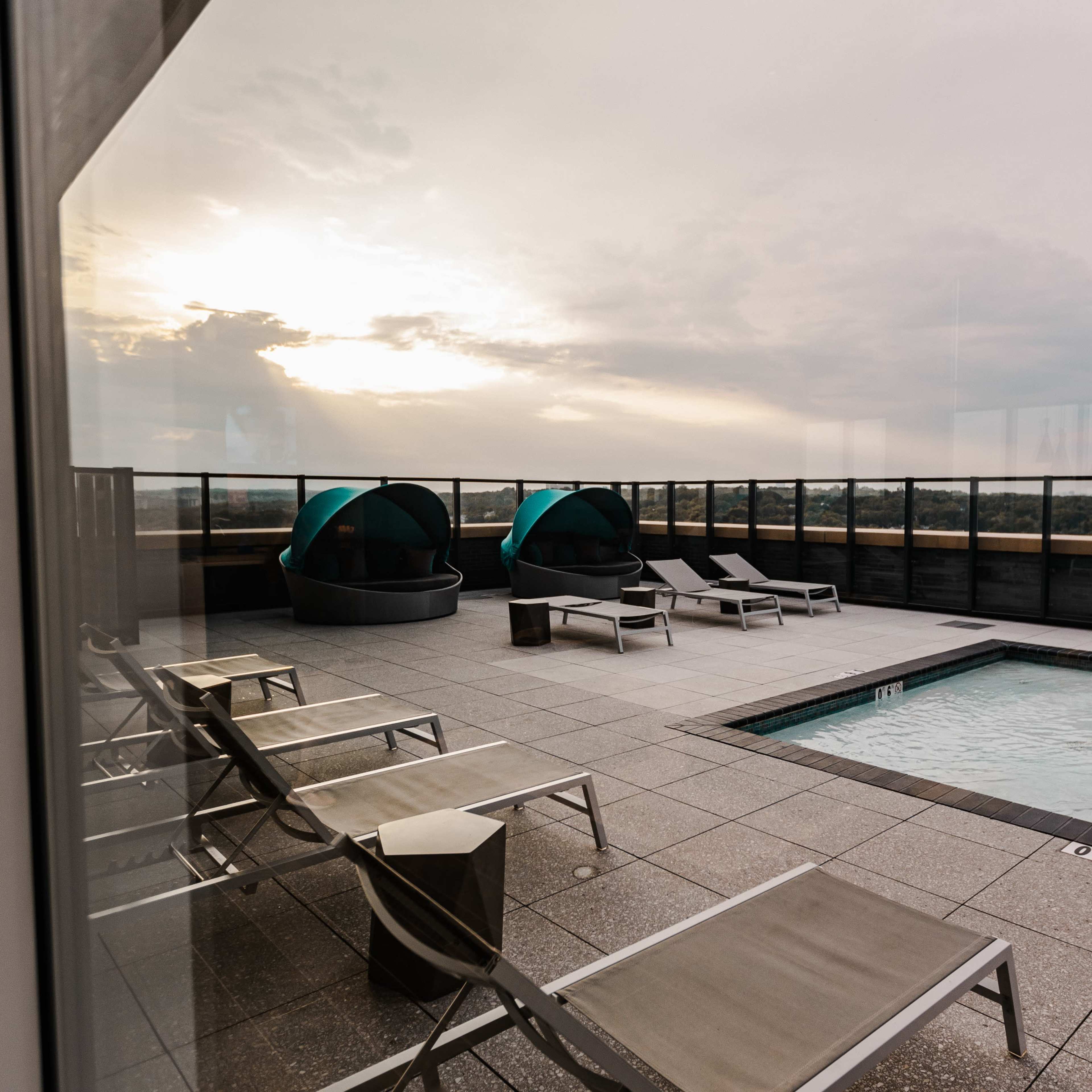 A rooftop pool area with lounge chairs, two cabanas, and a view of the horizon at sunset.