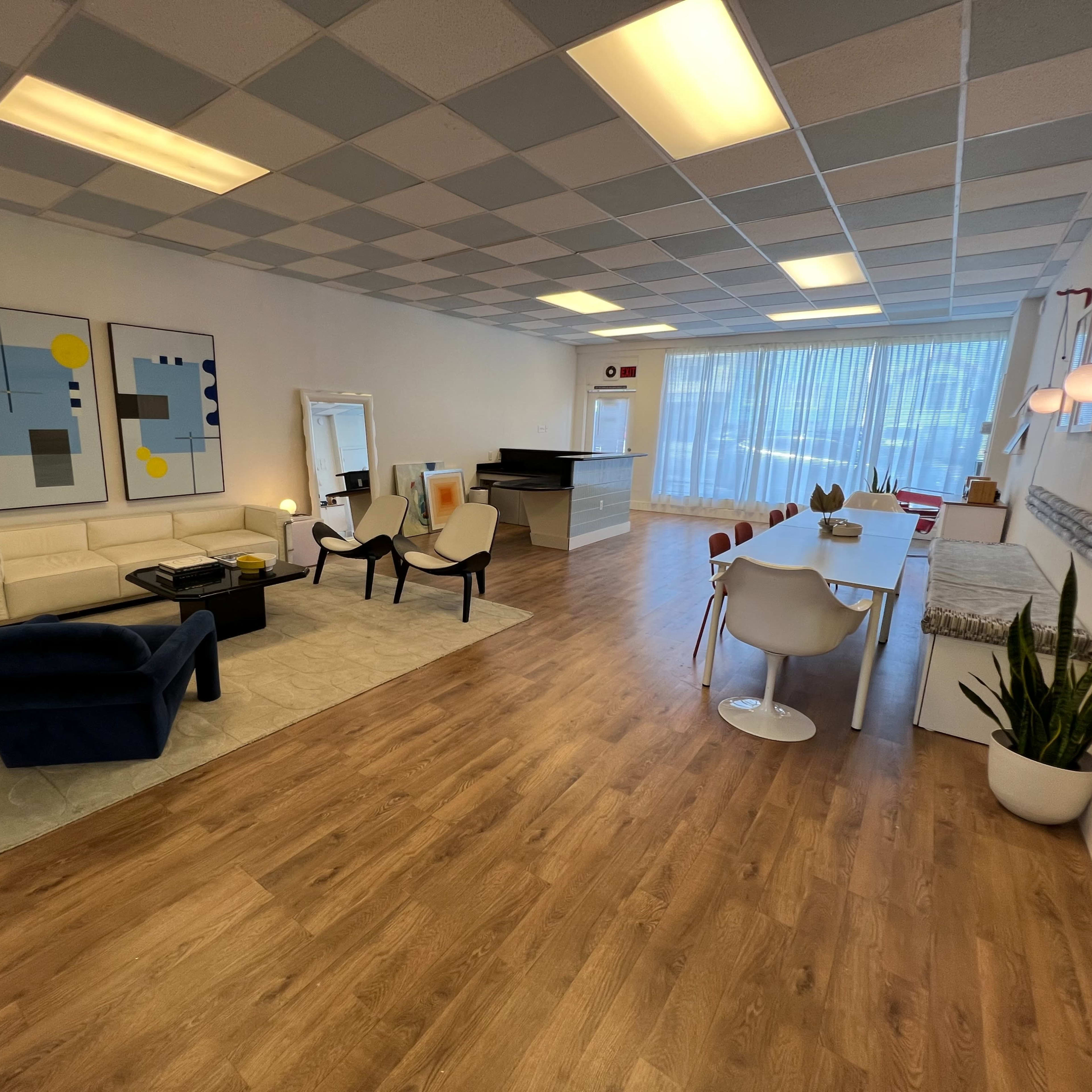 A spacious, well-lit room with a modern living area featuring a white sofa, chairs, and a dining table, along with large windows and artwork on the walls.
