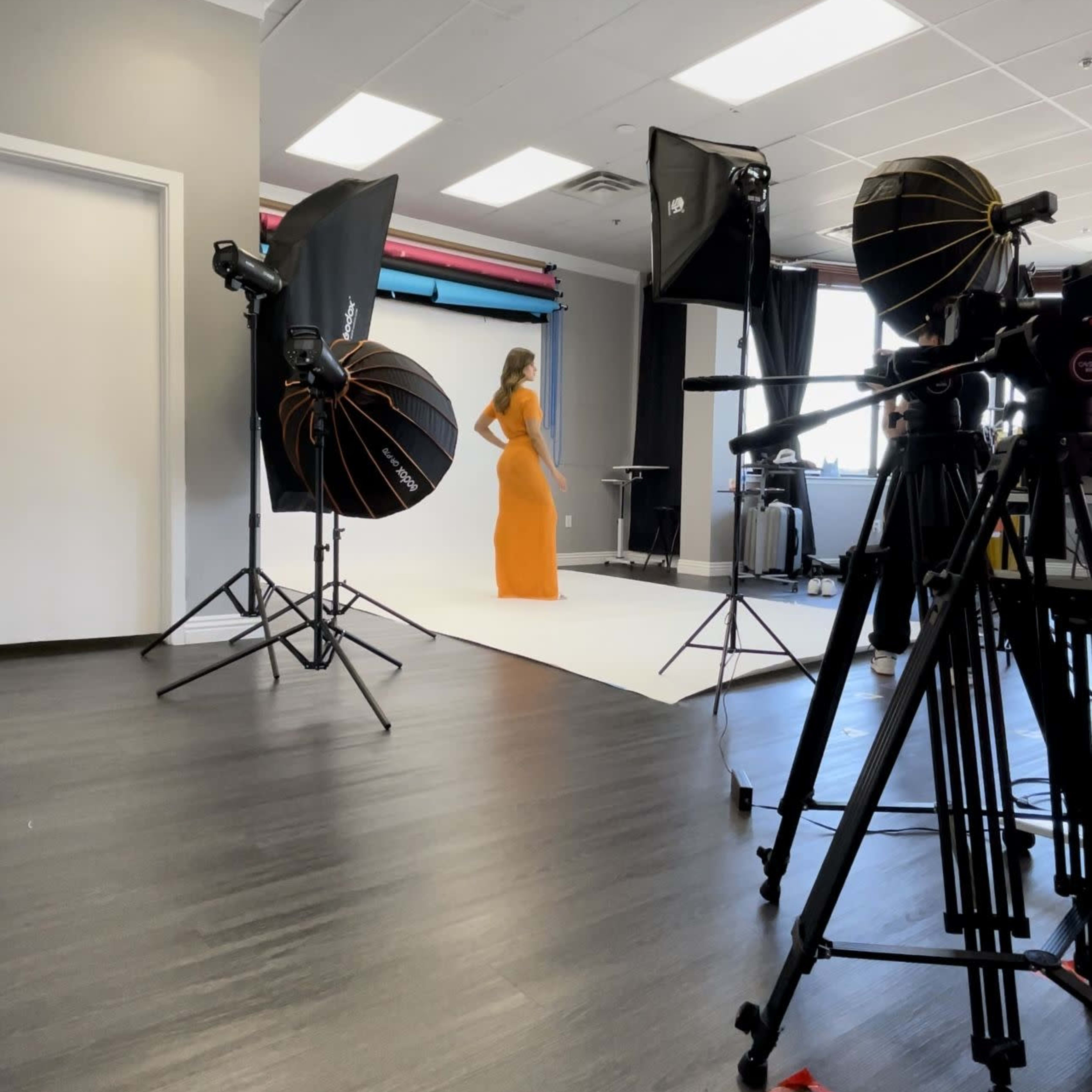 A model in an orange dress poses on a white backdrop in a photography studio equipped with lighting and cameras.