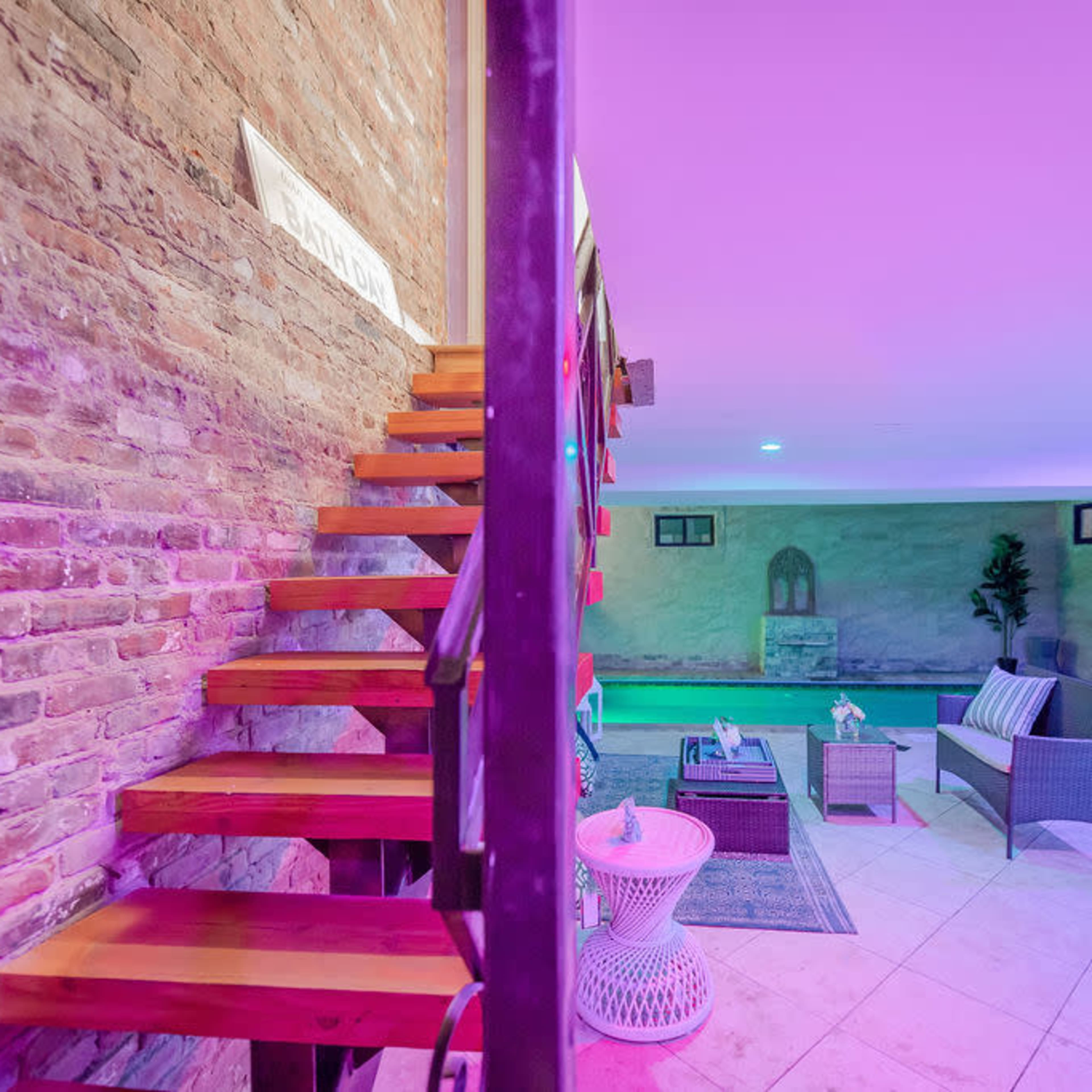 A staircase leads to a brightly lit basement lounge area furnished with seating and decorative elements against a brick wall.