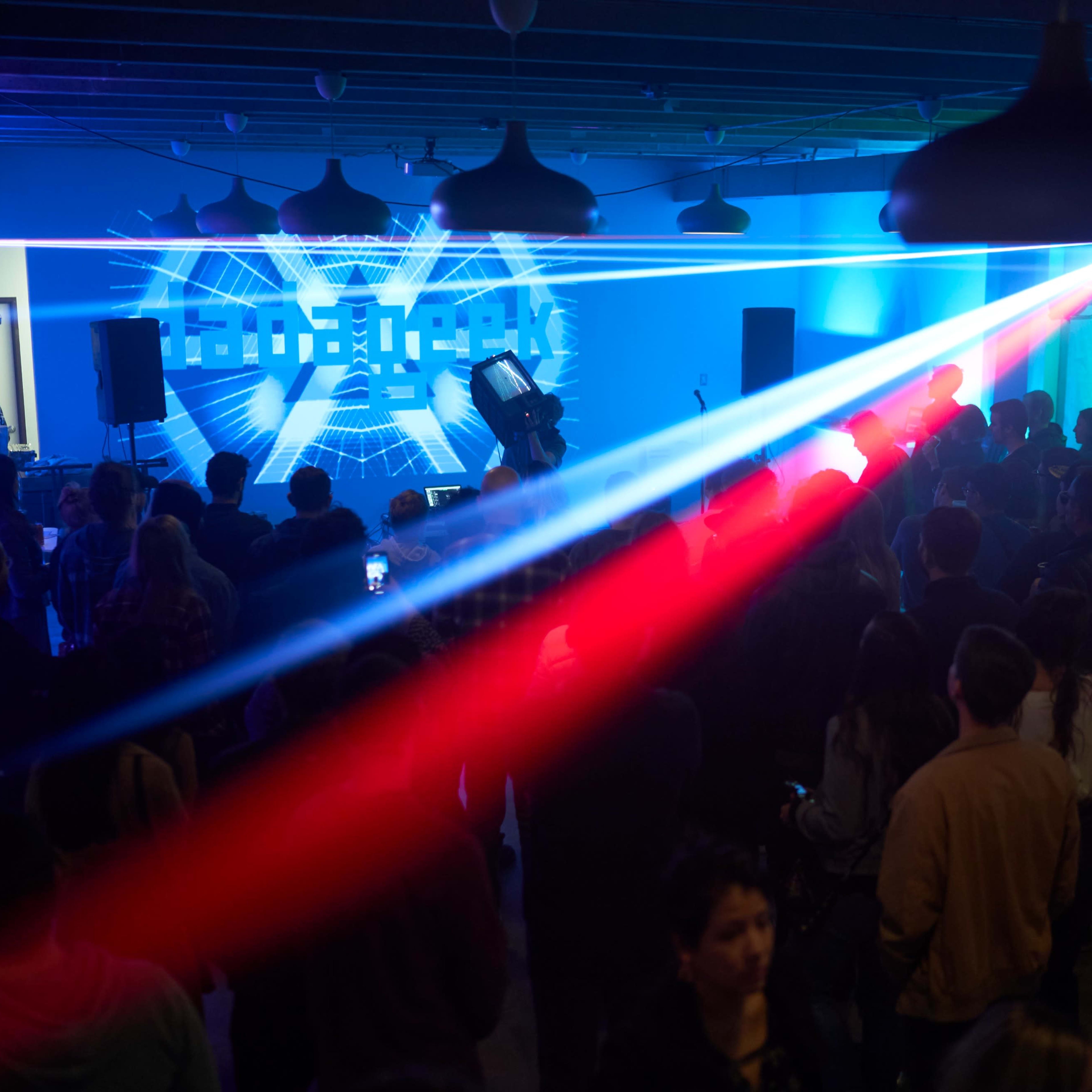 A crowd of people stands in a dimly lit venue with colorful laser lights projecting onto the wall during a live performance.