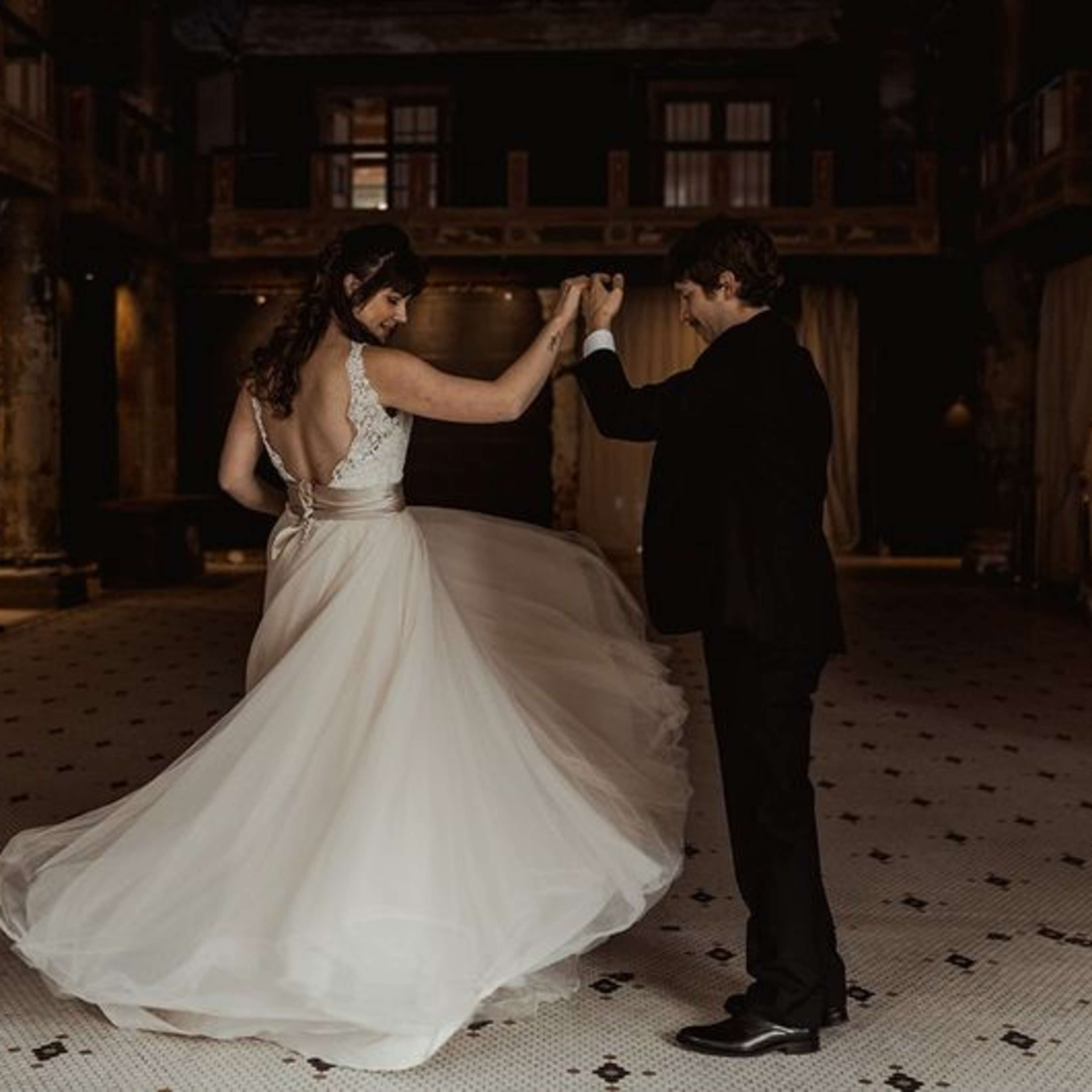 A couple dances in a spacious, dimly lit venue with a patterned tiled floor and exposed structural elements.
