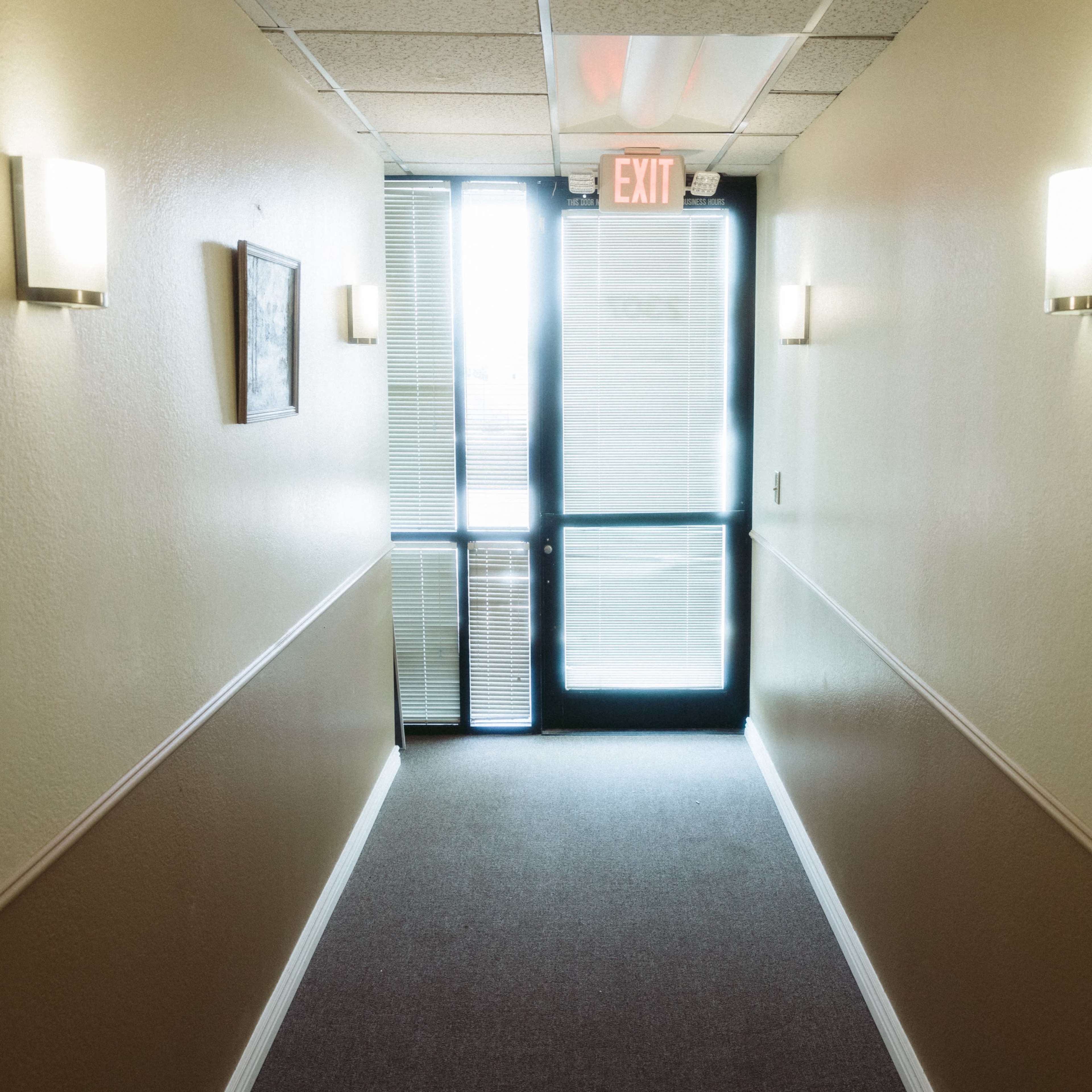 A brightly lit hallway leads to a glass exit door, with wall sconces illuminating the beige walls and a framed picture on the left.