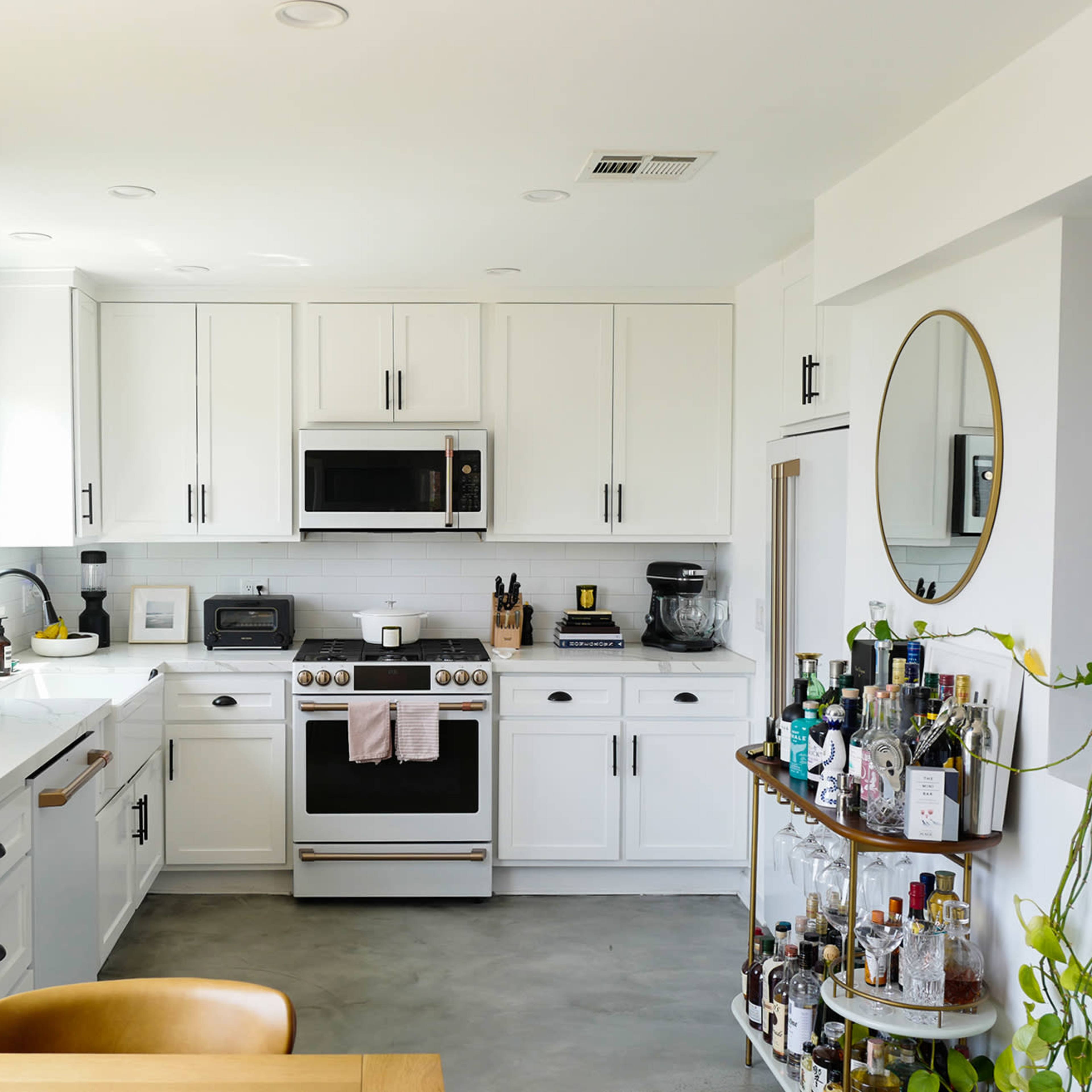 A modern kitchen features white cabinetry, stainless steel appliances, a countertop with coffee equipment, and a circular mirror above a bar cart filled with various bottles.
