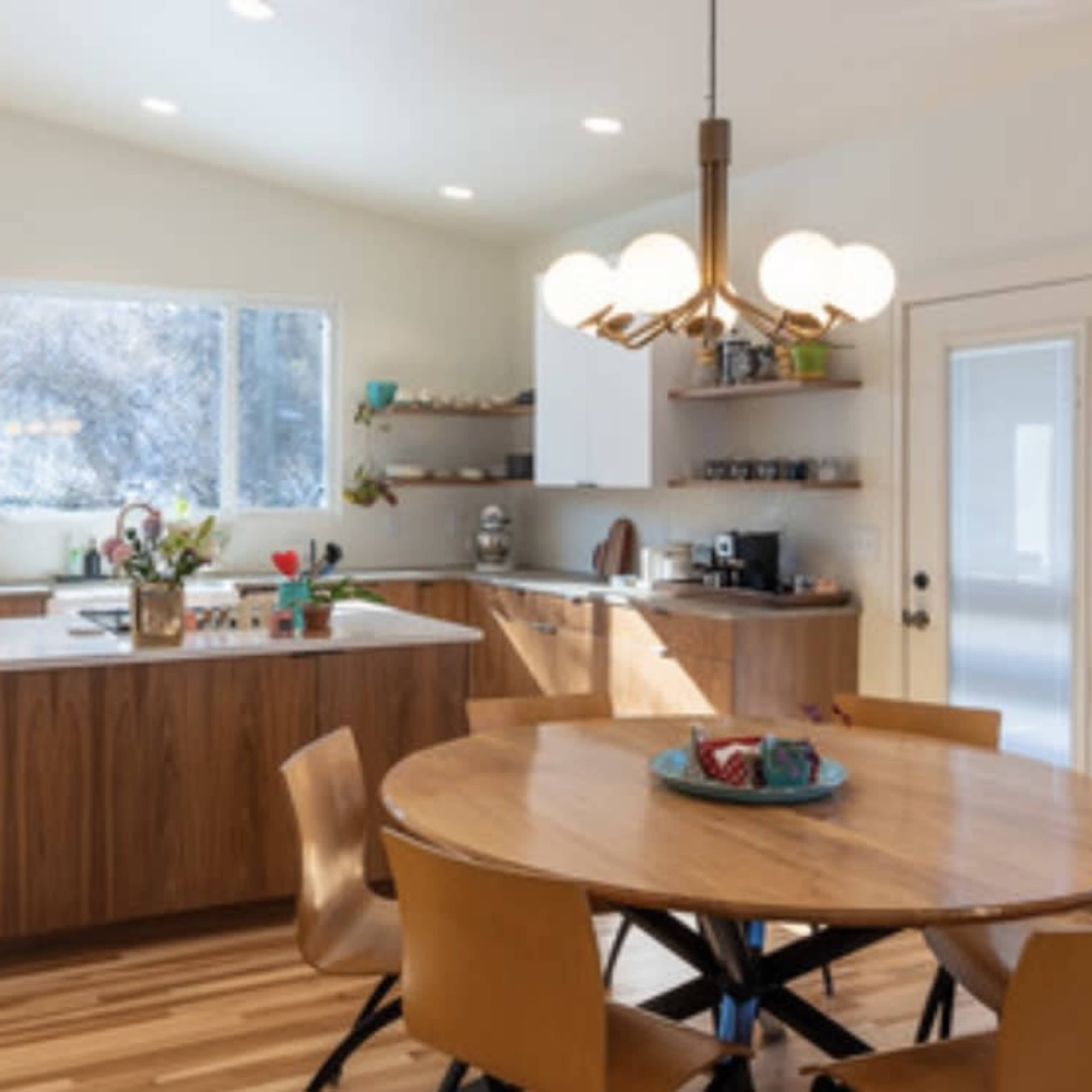 A modern kitchen features wooden cabinetry, a central round dining table, and large windows that let in natural light.