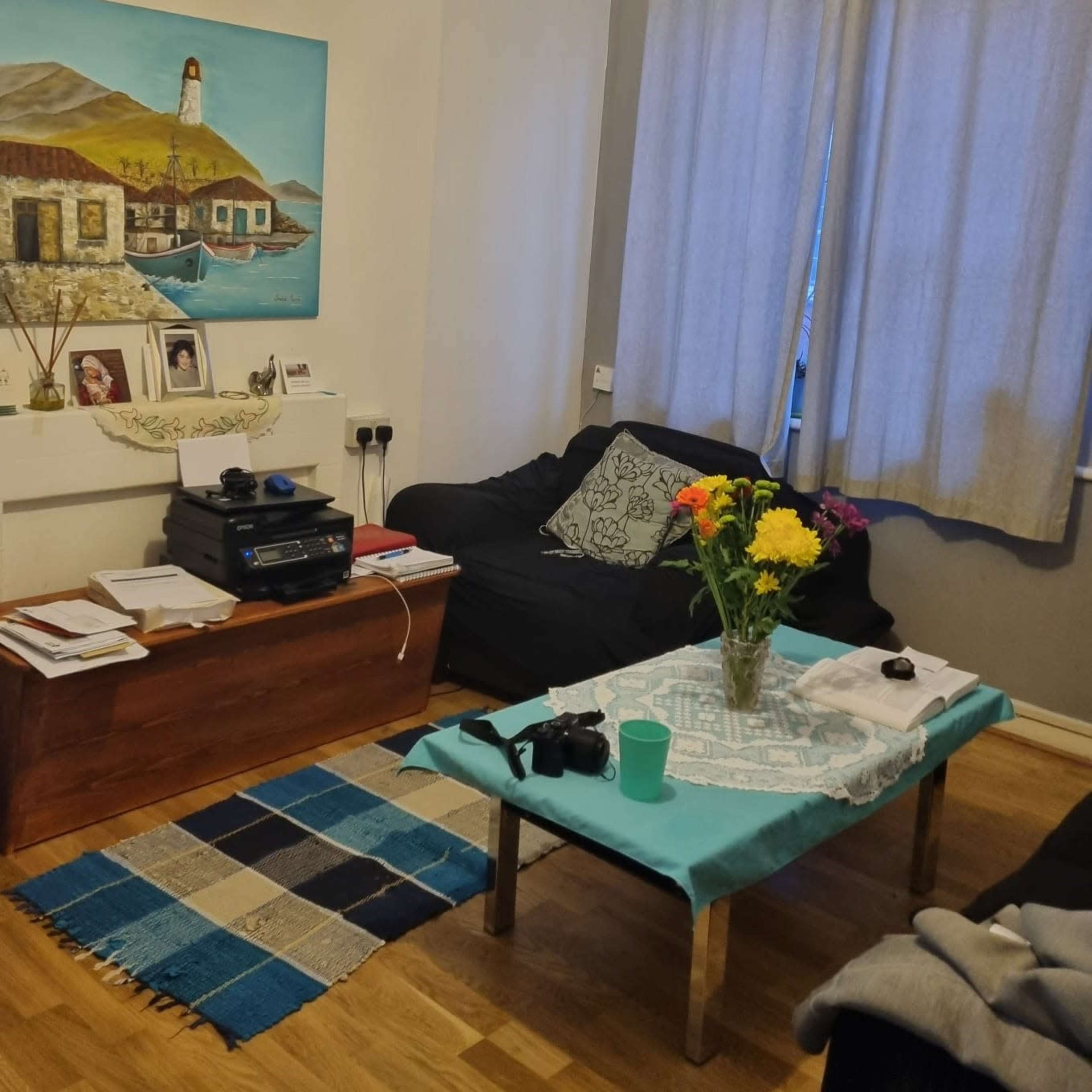 A cozy living room with a blue and white theme, featuring a wooden coffee table, a dark couch, a colorful painting on the wall, and a bouquet of flowers in a vase.