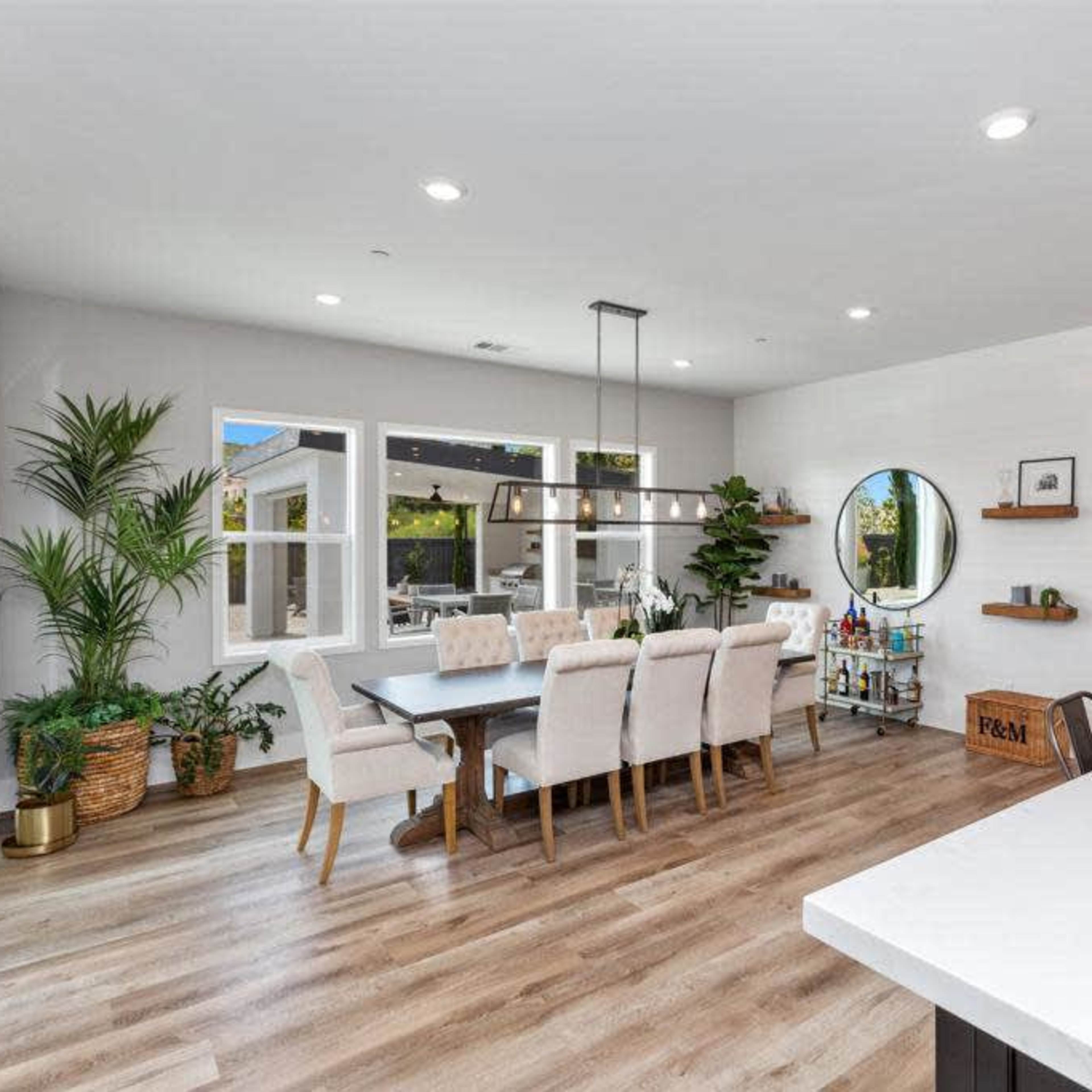 A modern dining area with a large table surrounded by upholstered chairs, large windows, and a circular mirror on the wall.