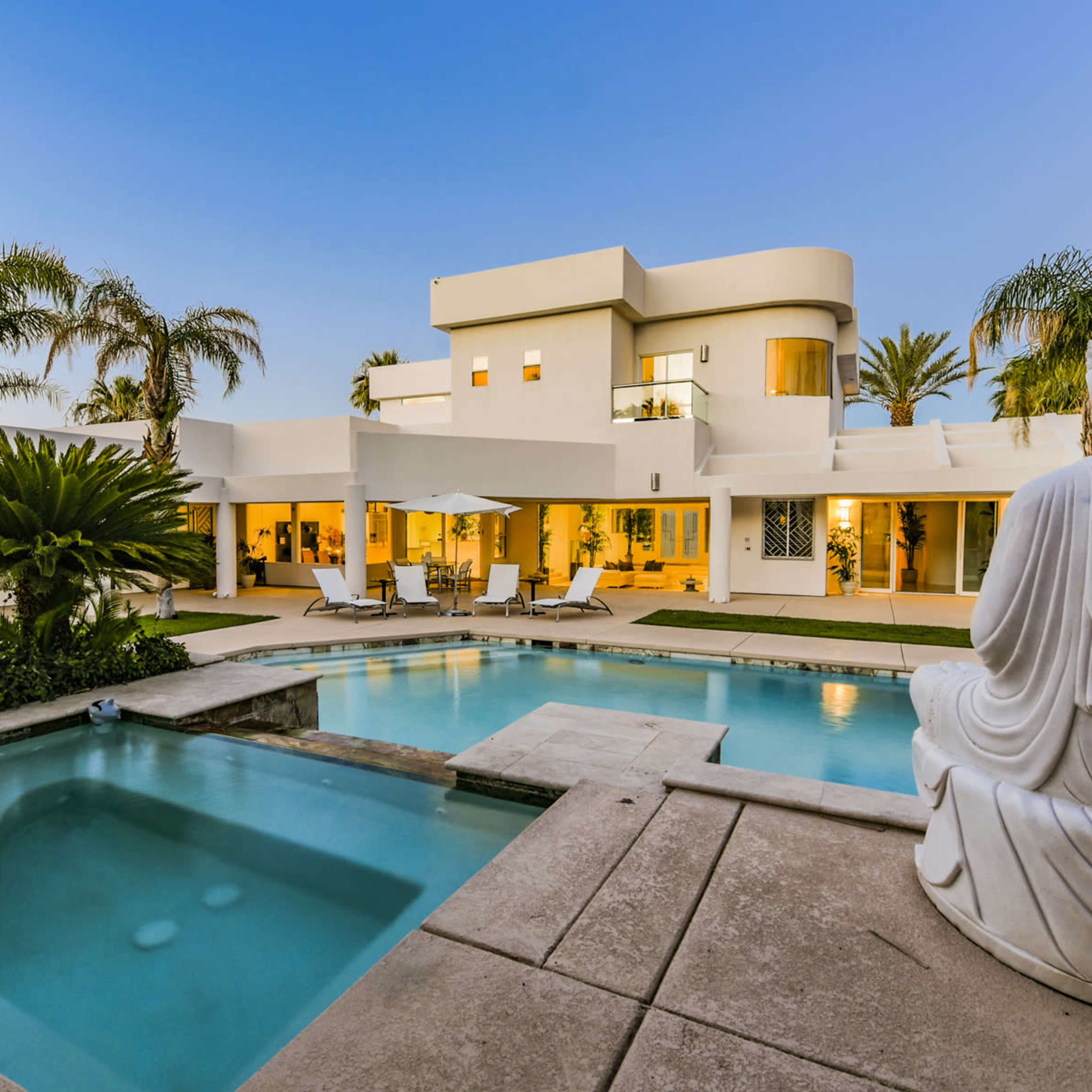 A modern villa features a swimming pool surrounded by palm trees and a statue of Buddha in the foreground.