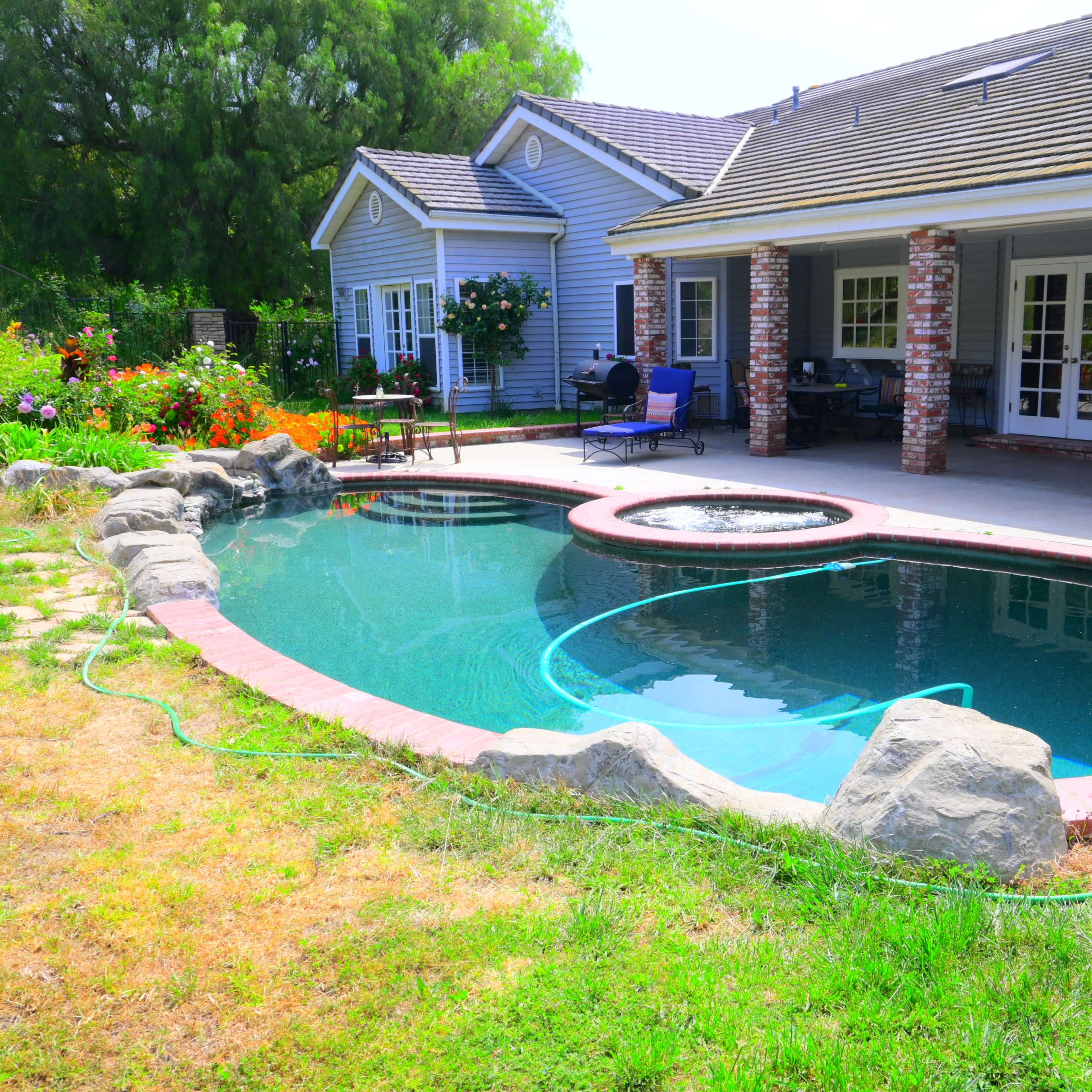A backyard with a landscaped area featuring a swimming pool surrounded by rocks and grass, adjacent to a house with a porch.