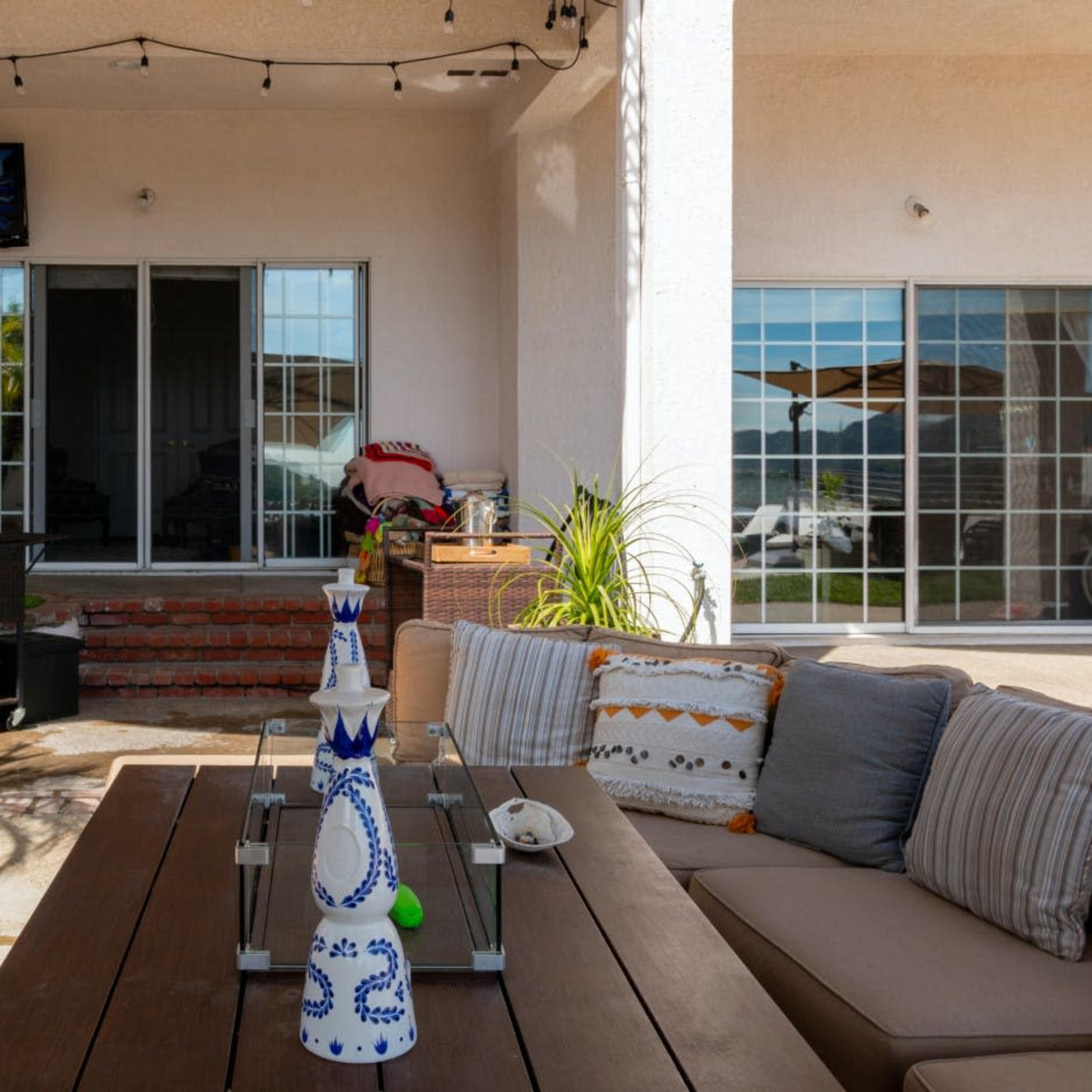 A patio area features a wooden table with a decorative item, surrounded by outdoor seating and a grill nearby, with large windows and a television mounted on the wall.