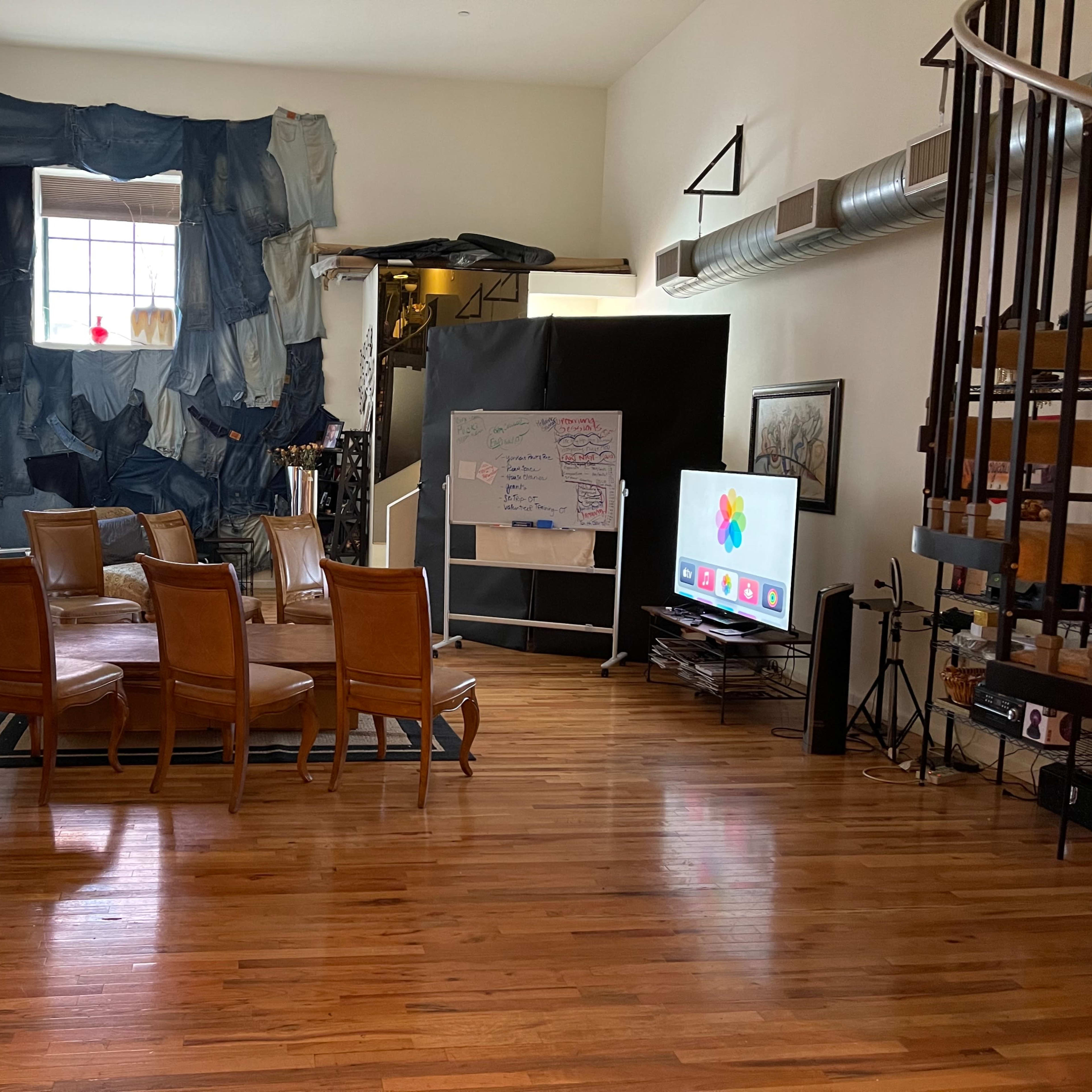 A spacious room features wooden flooring, a circular table surrounded by six chairs, a television, and a wall decorated with fabric.