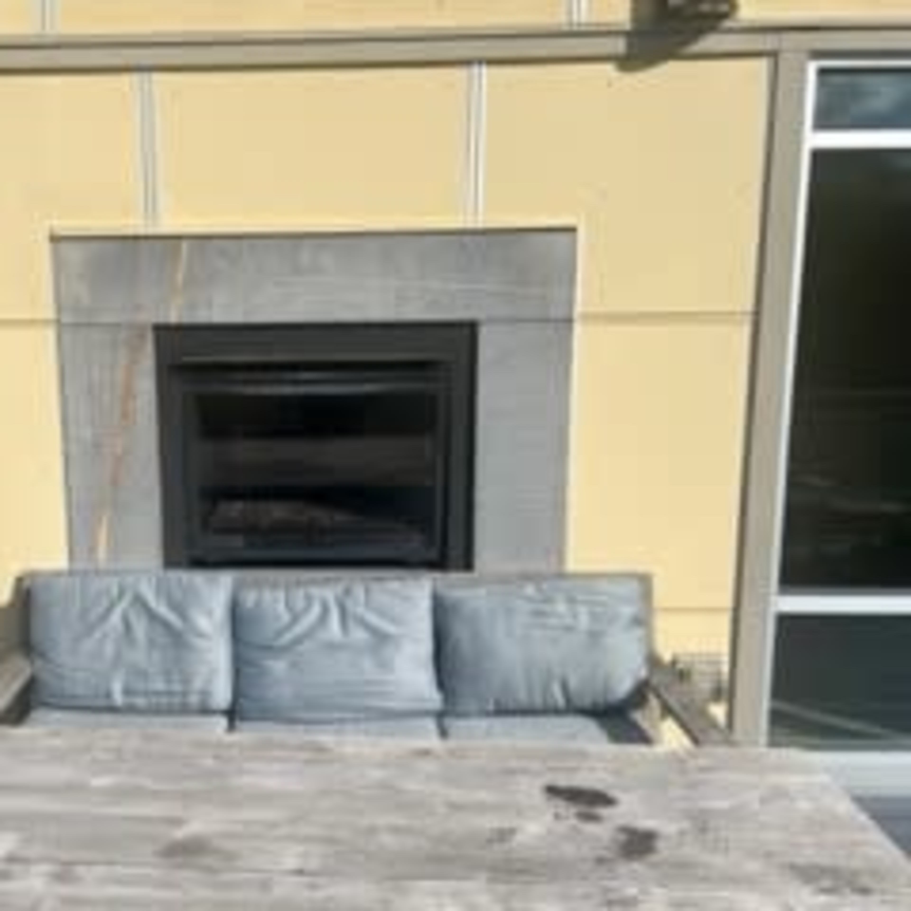 A gray sofa is positioned in front of a modern fireplace on a patio with large windows.