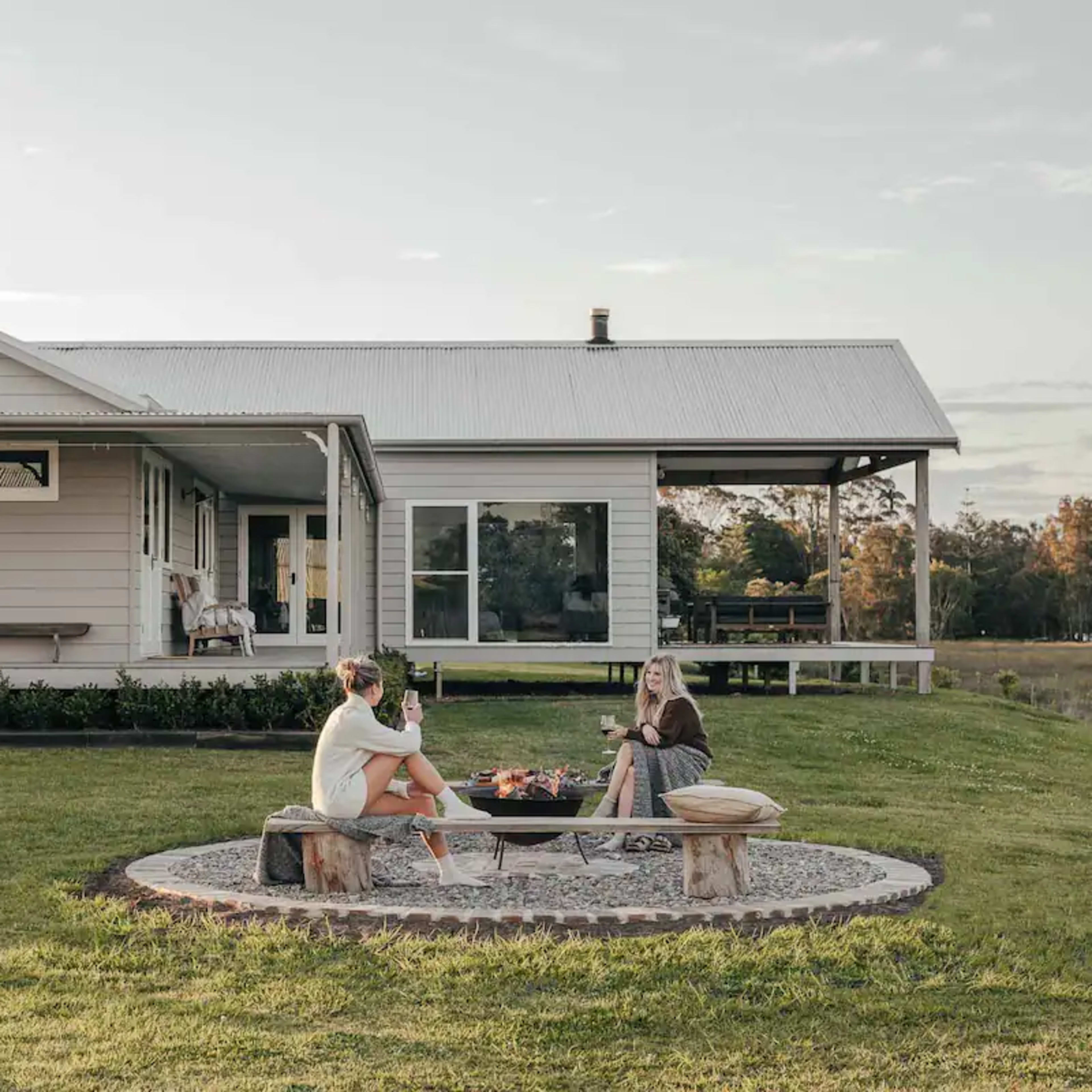 Two women sit around a stone fire pit in the yard of a light-colored house, enjoying drinks and snacks.