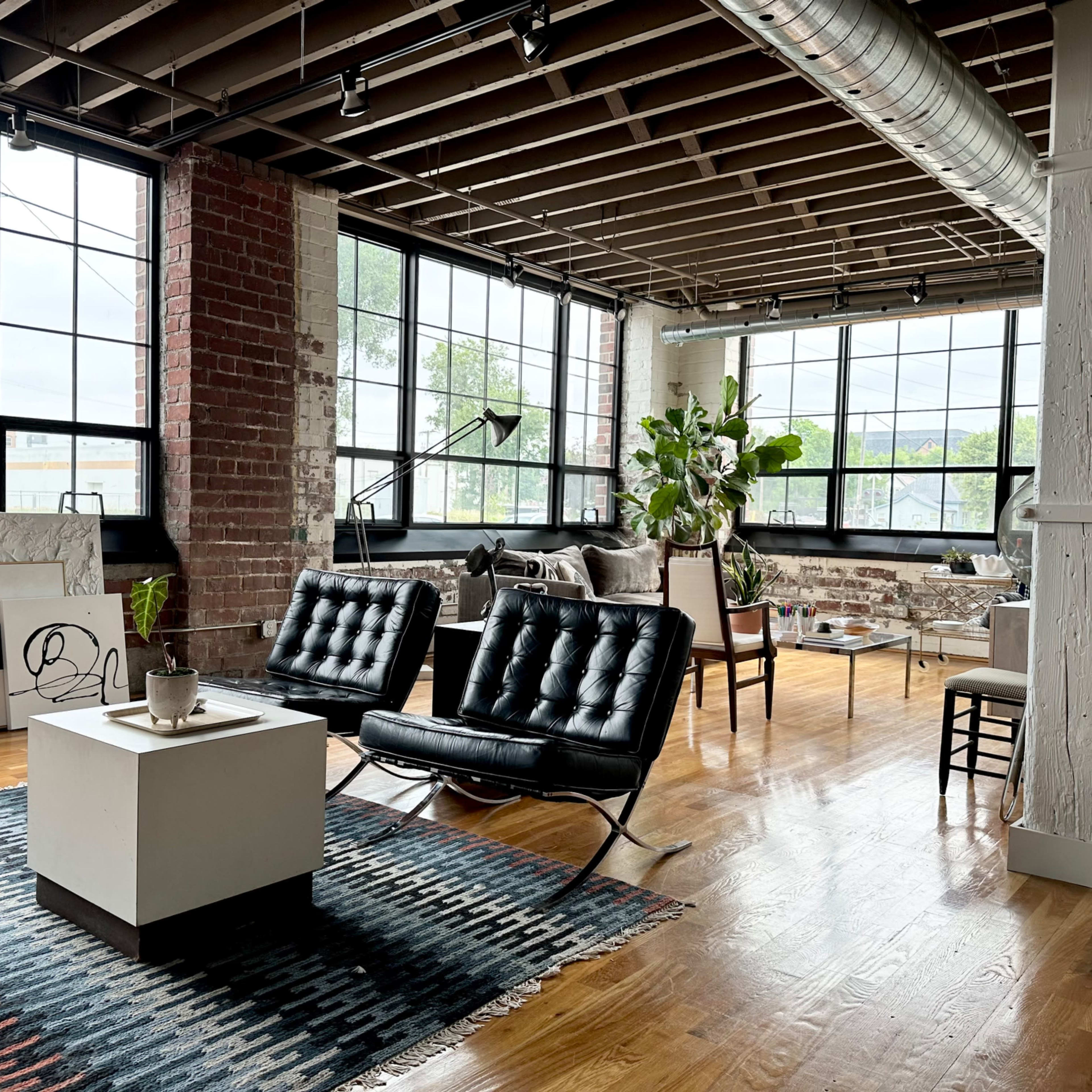 A modern loft-style living room with large windows, industrial brick walls, and contemporary furniture arranged around a central coffee table.