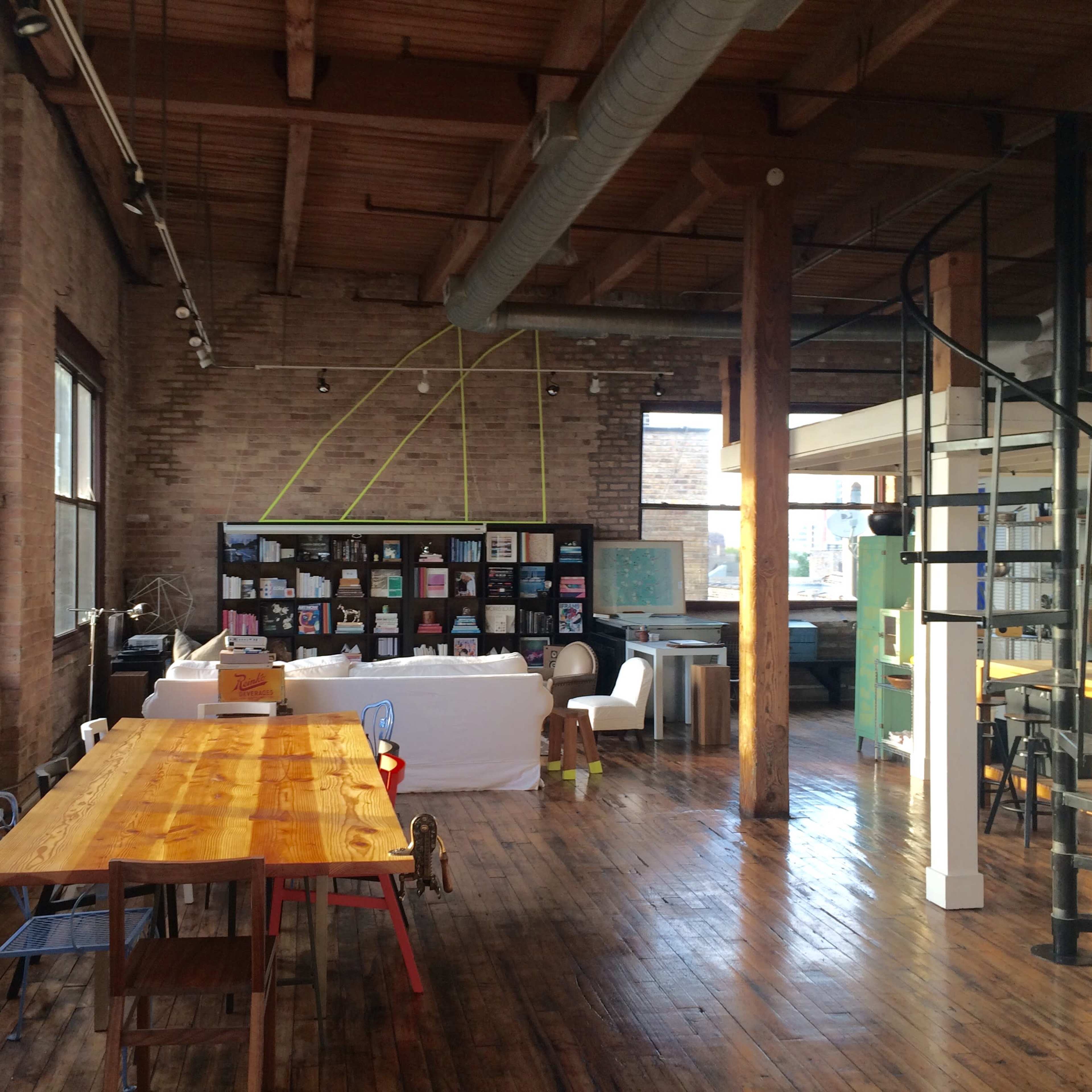 A spacious, industrial-style room with large windows, wooden flooring, and a long dining table, along with various furniture pieces and a spiral staircase.